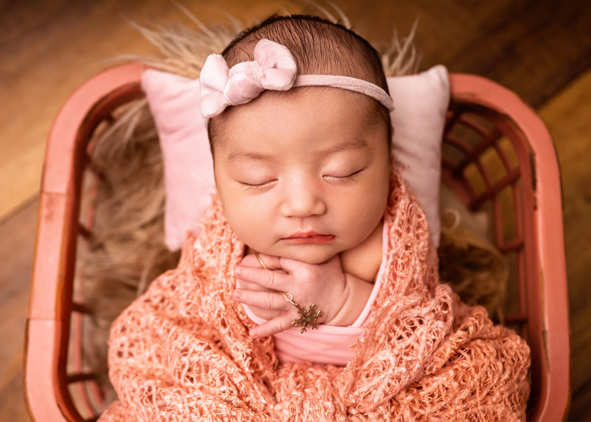 A sleeping baby girl with a pink headband and bow, wrapped in a peach-colored knitted blanket, lying on a pink pillow inside a pink basket with a furry lining.