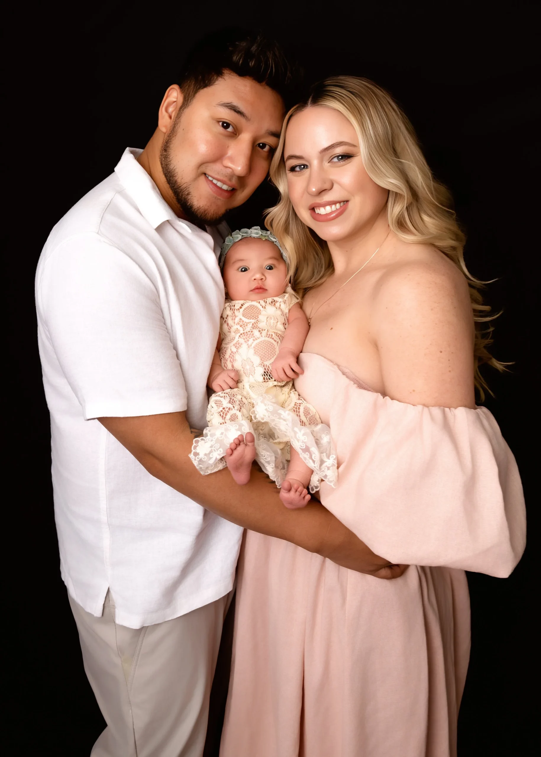 A family portrait with a man, woman, and baby against a black background. The man has short dark hair and a beard, wearing a white shirt. The woman has long blonde hair and is wearing a light pink off-the-shoulder dress. The baby is dressed in a lace