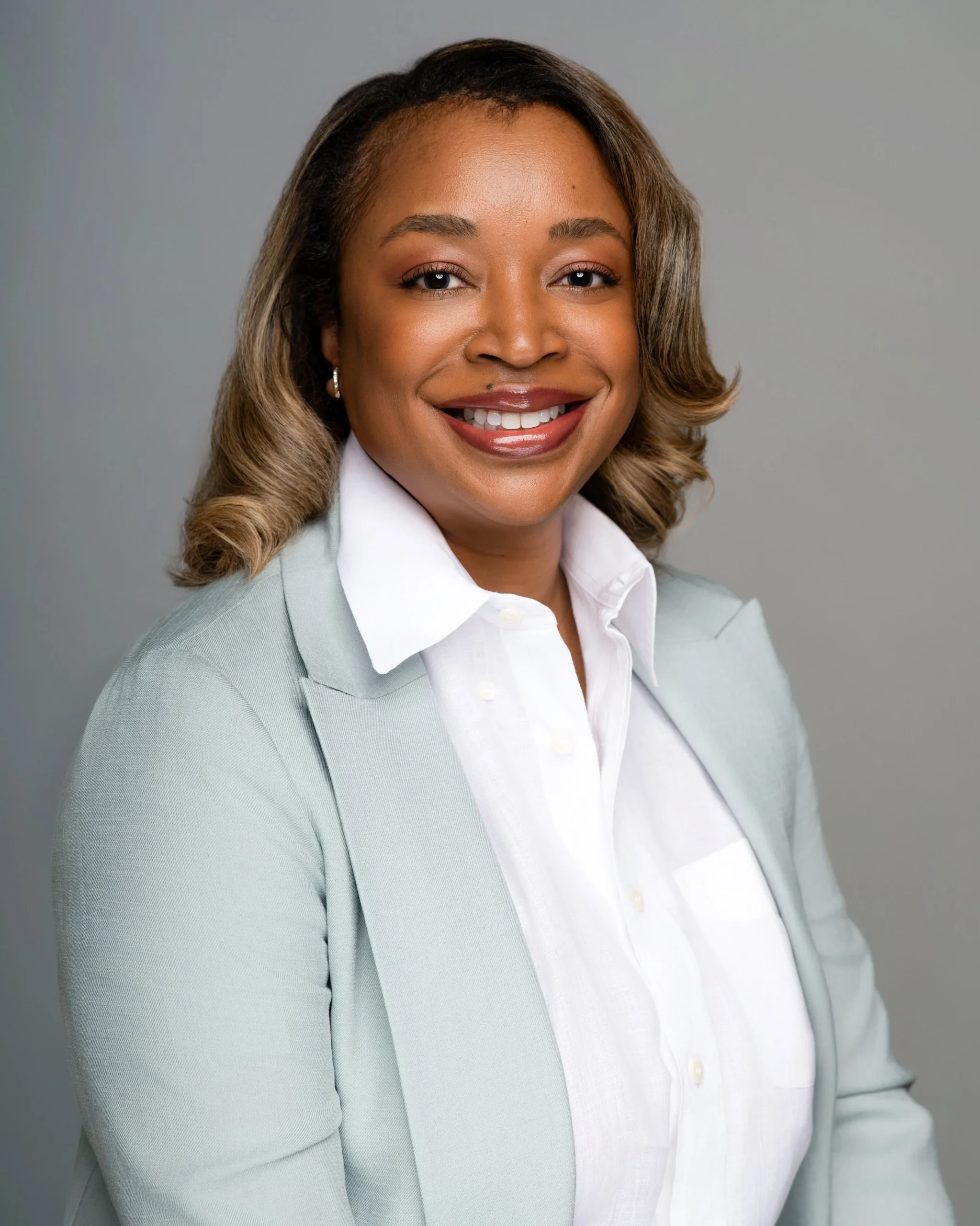 A professional woman smiling, wearing a white shirt and light gray blazer against a plain gray background.