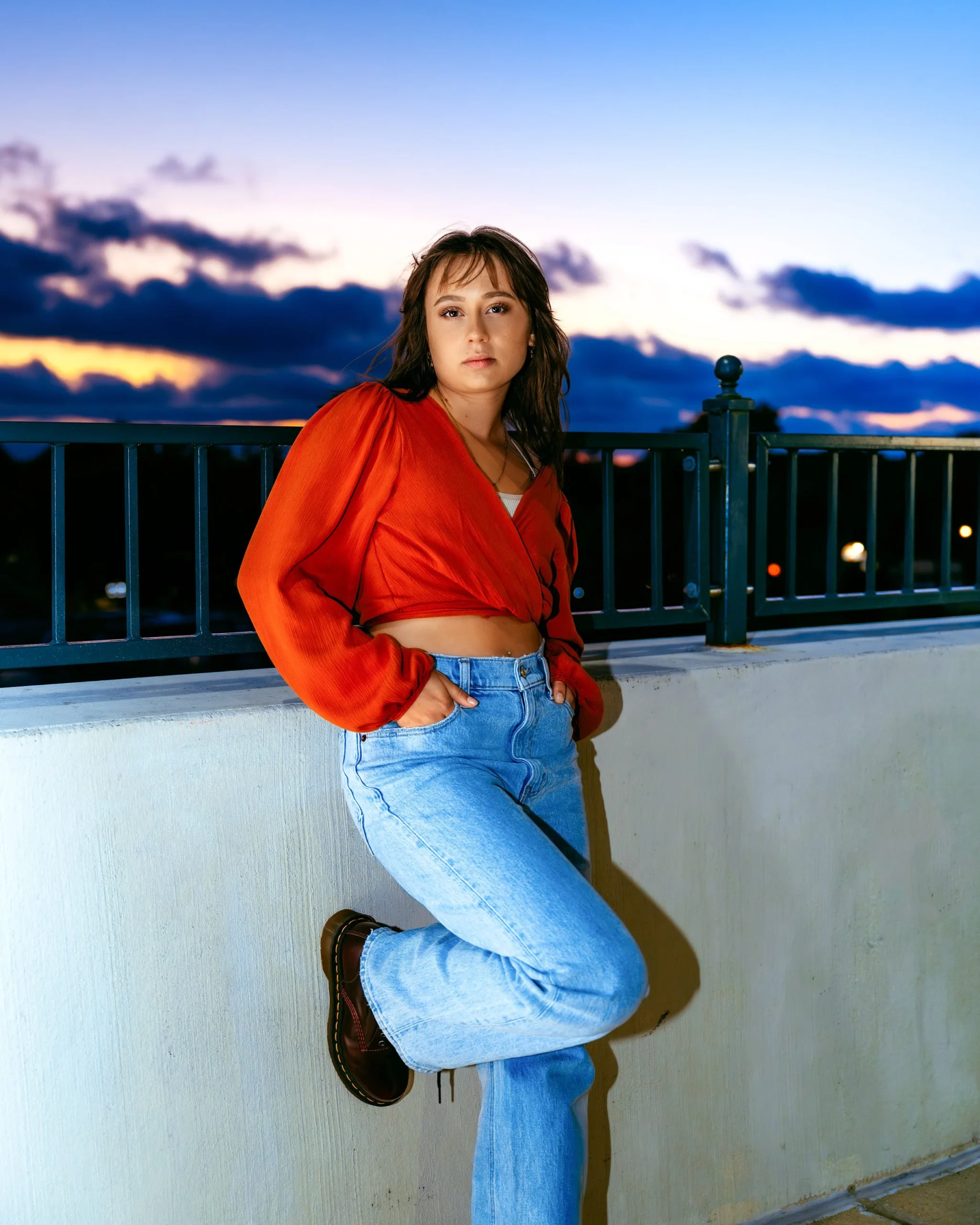 Person in orange top and jeans leaning against a railing at sunset.