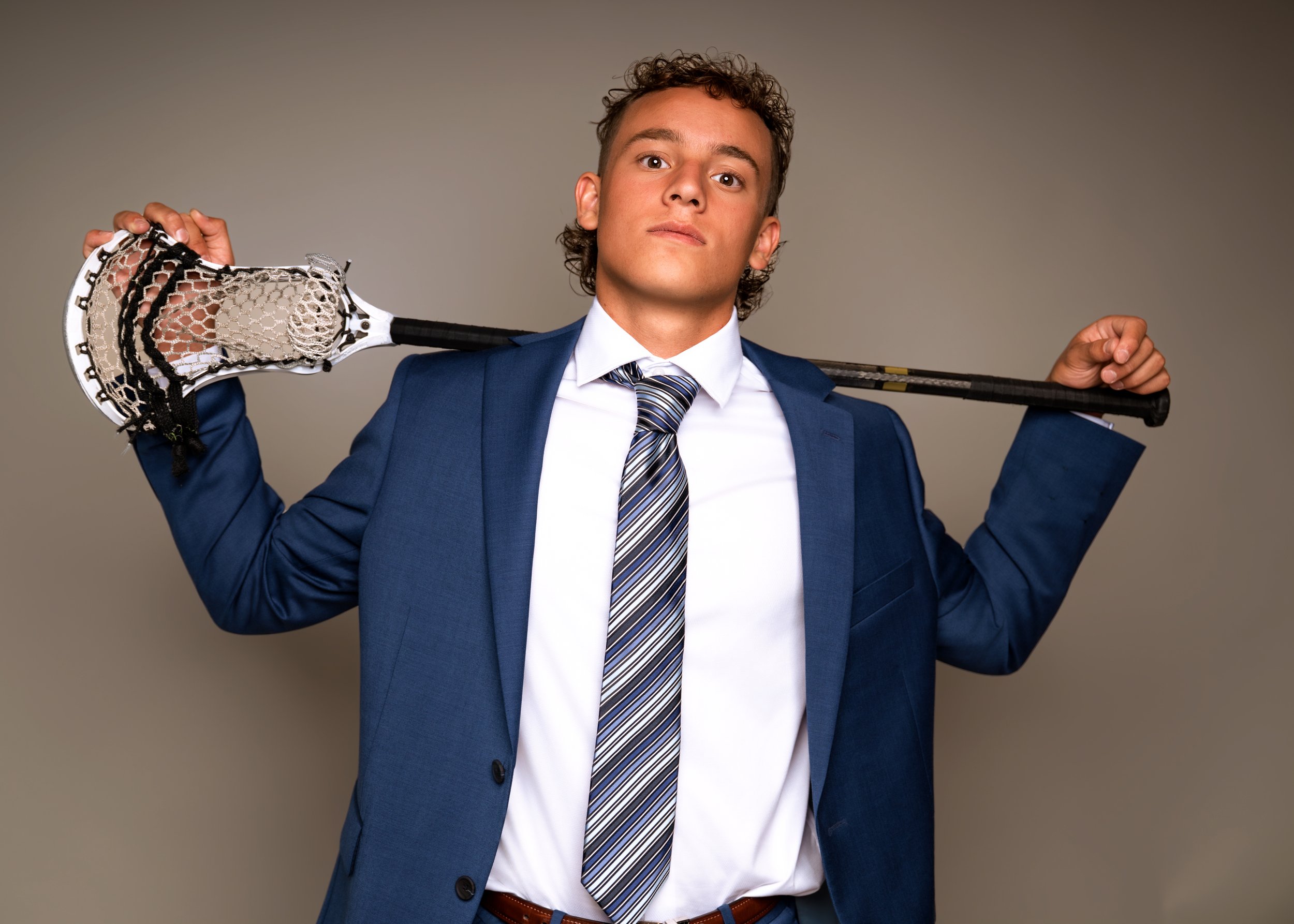 Young man in a blue suit, white shirt, and striped tie holding a hockey stick over his shoulders, posing confidently against a plain background.