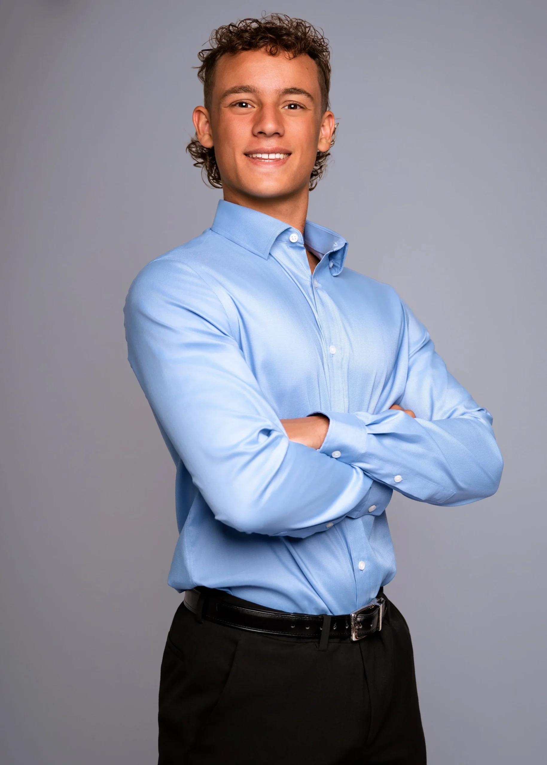 Portrait of a young man with curly hair, wearing a light blue dress shirt and black pants, standing with arms crossed against a neutral background.