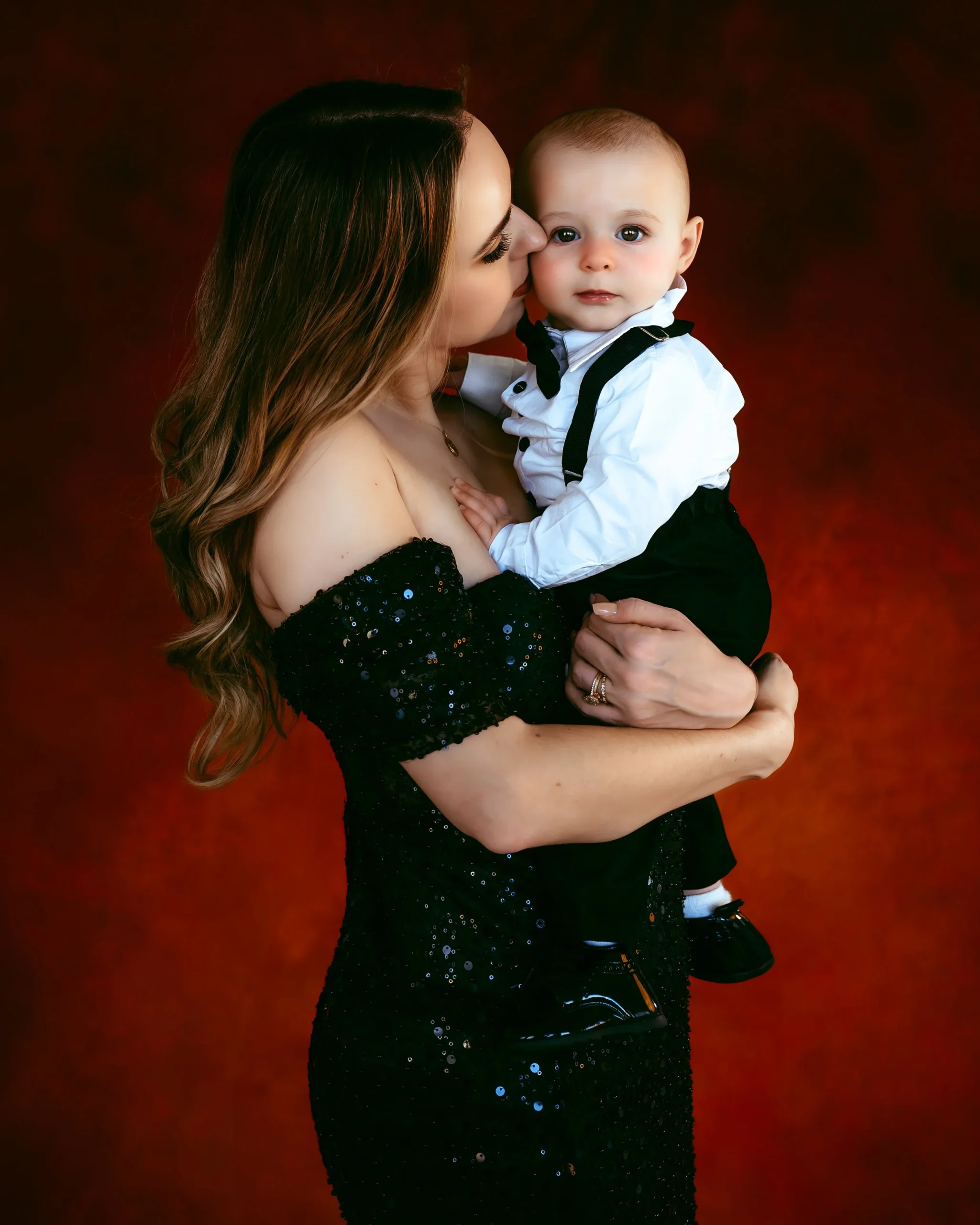 Woman in a black sequin dress holds a baby in formal attire against a red background.