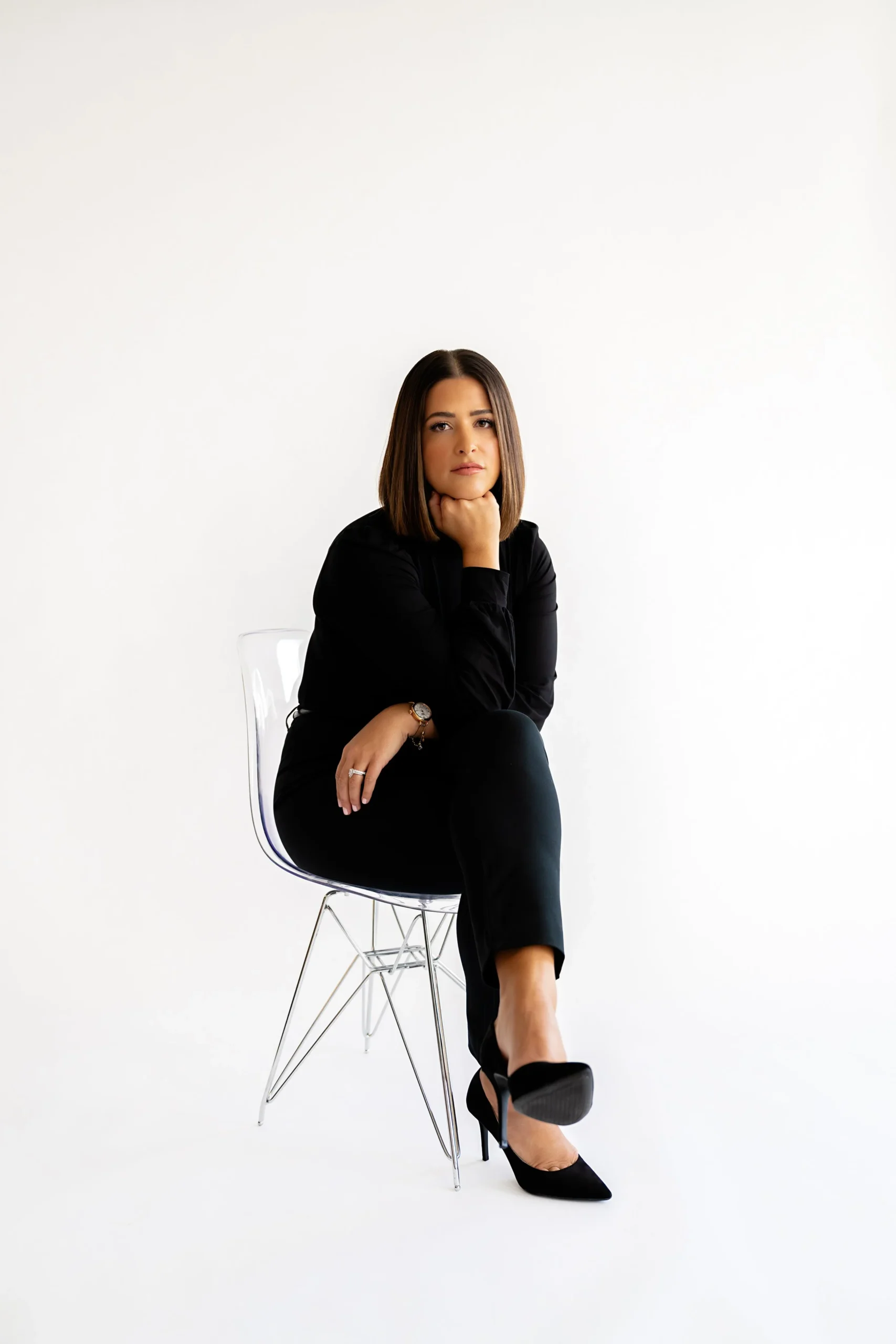 A woman in a black outfit sitting on a clear modern chair against a white background, with her chin resting on her hand.