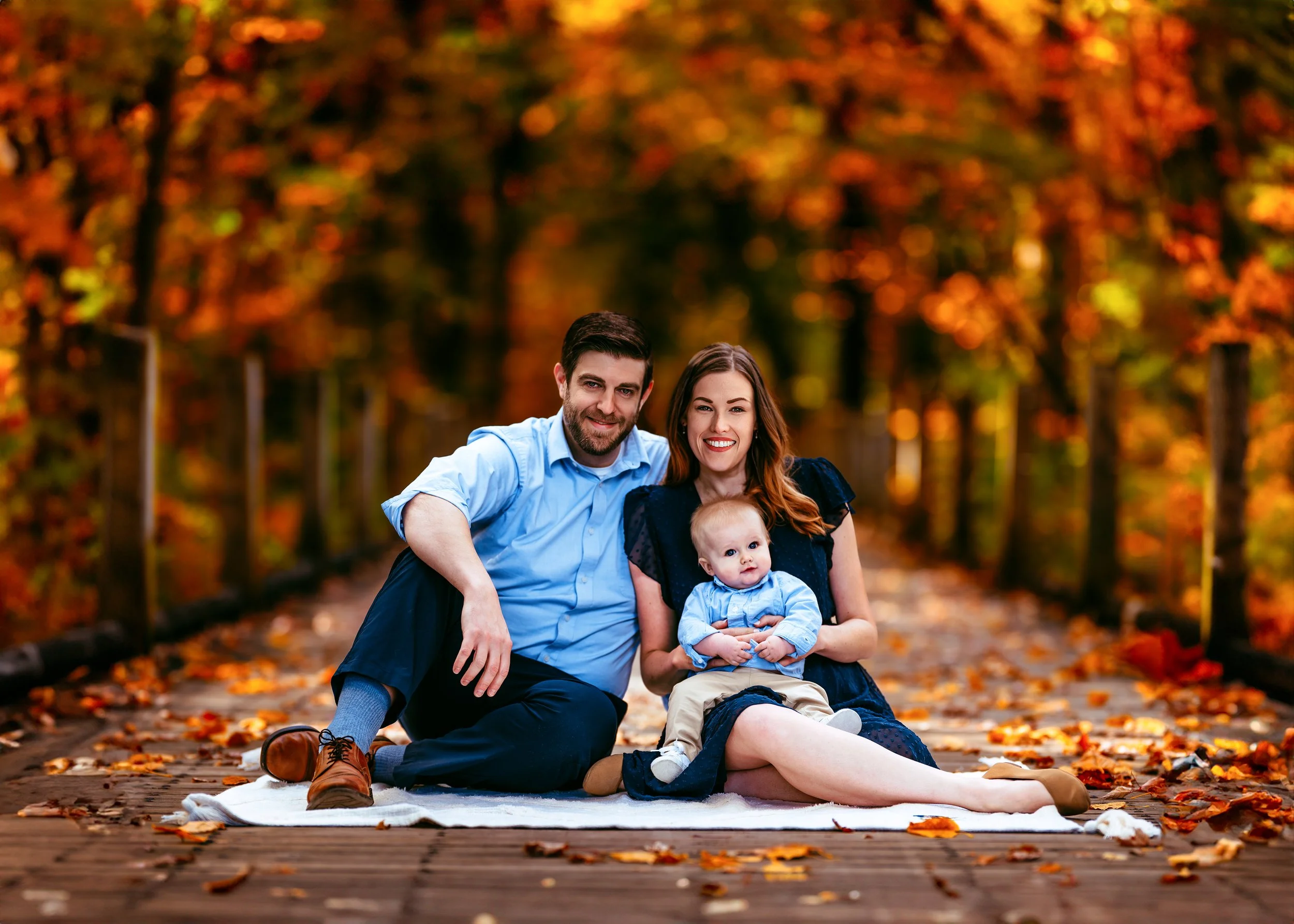 Family sitting on a blanket in a park during fall, surrounded by autumn leaves.