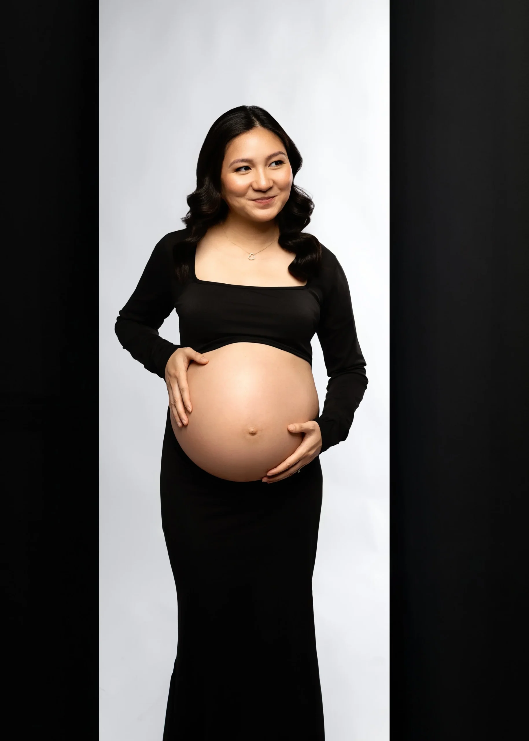 A pregnant woman with long black hair, smiling, wearing a black dress and a black long-sleeve top, standing against a gray and black background, holding her belly with both hands.