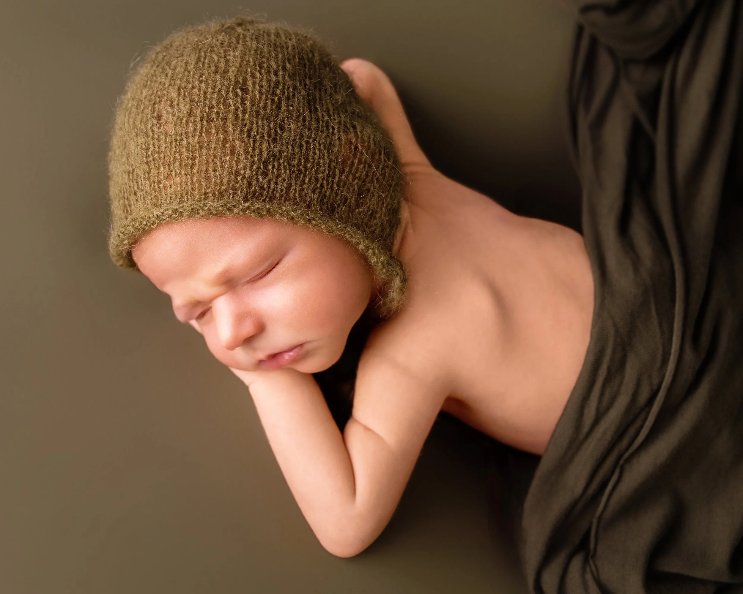A newborn baby sleeping peacefully with a brown knit hat, resting its head on its arm and covered partly with a black blanket.