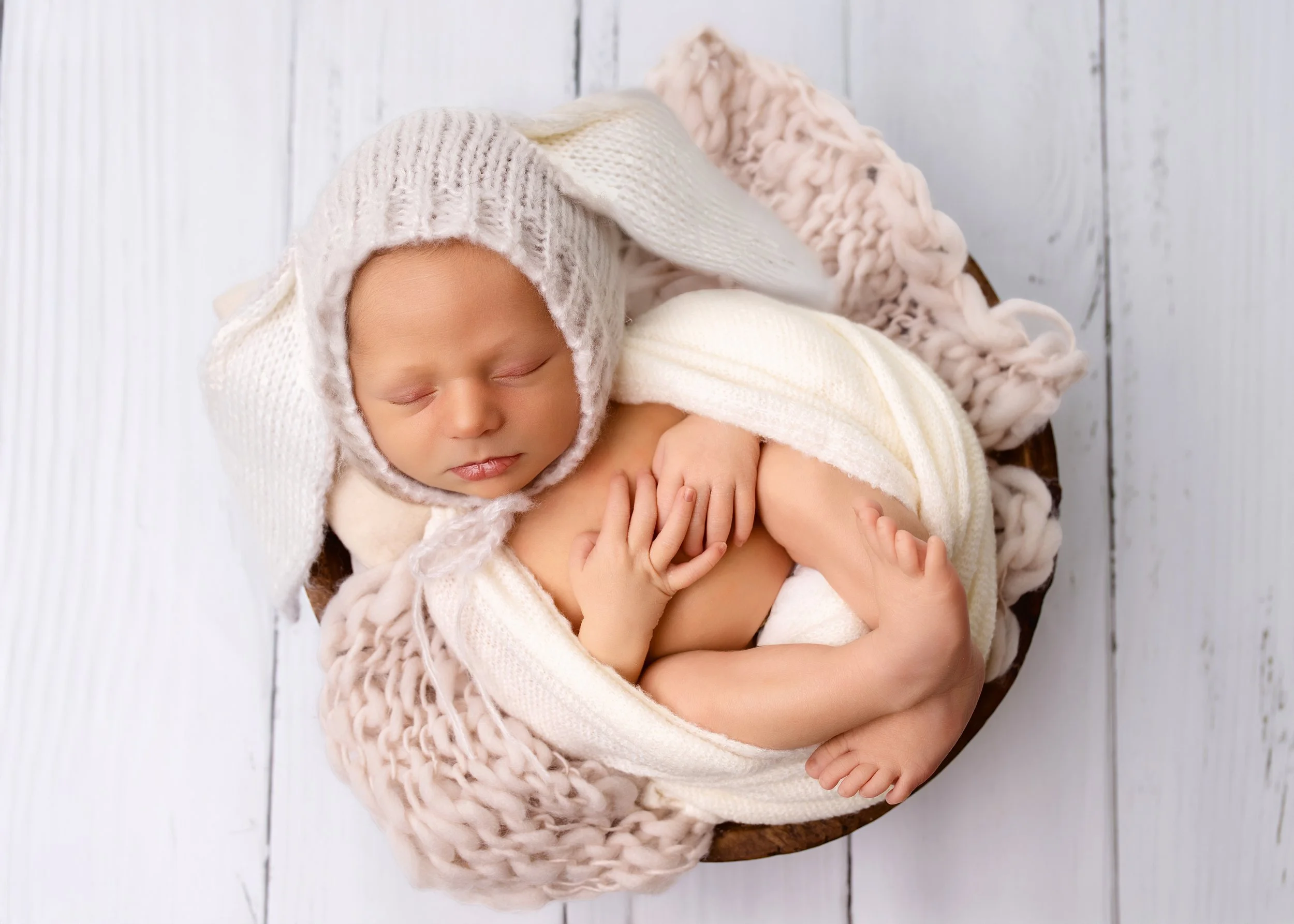 A sleeping newborn baby wrapped in cream-colored blankets, wearing a knitted white hat, lying in a basket on a white wooden floor.