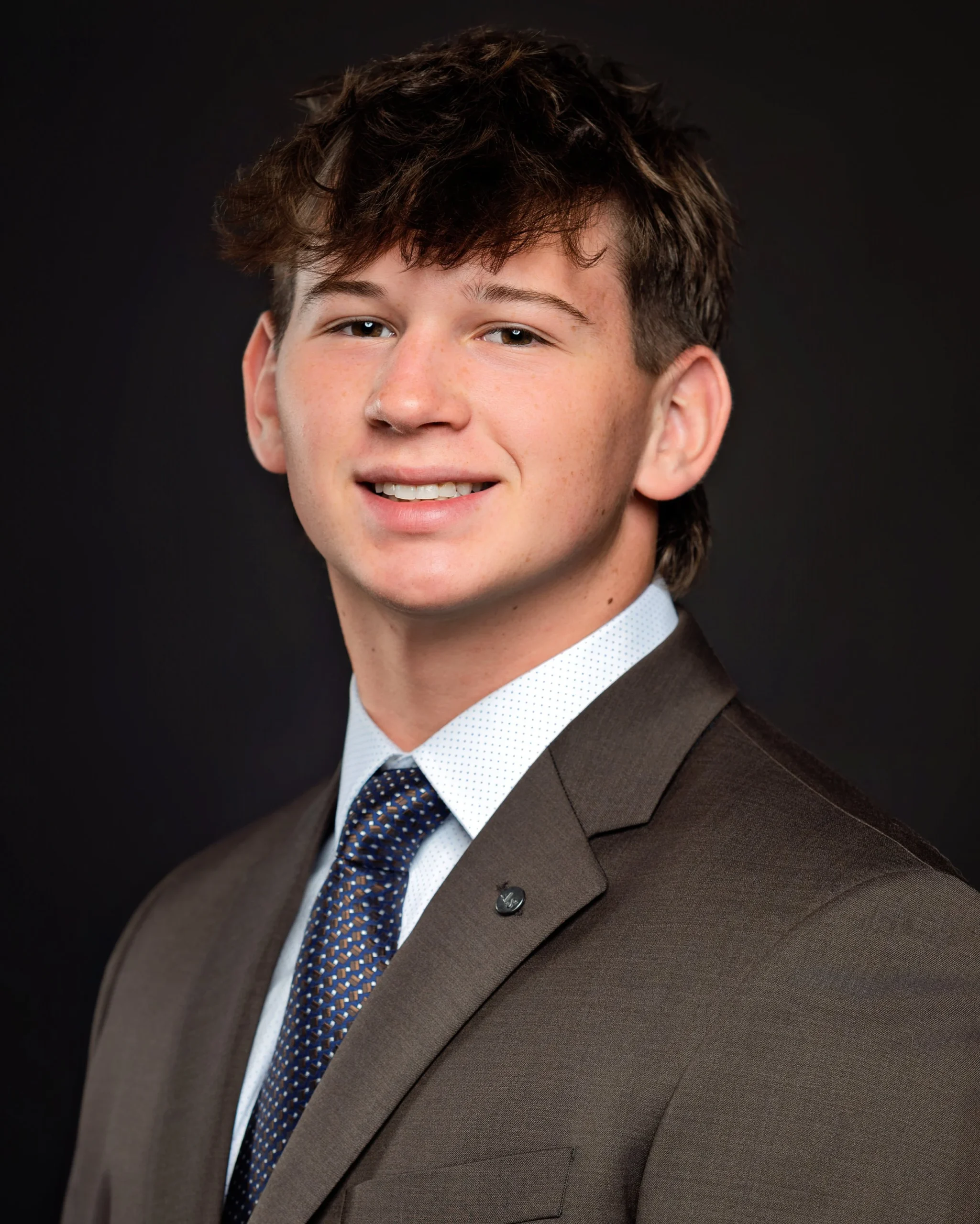 Professional headshot of a young man with brown hair, wearing a suit and tie, smiling against a dark background.