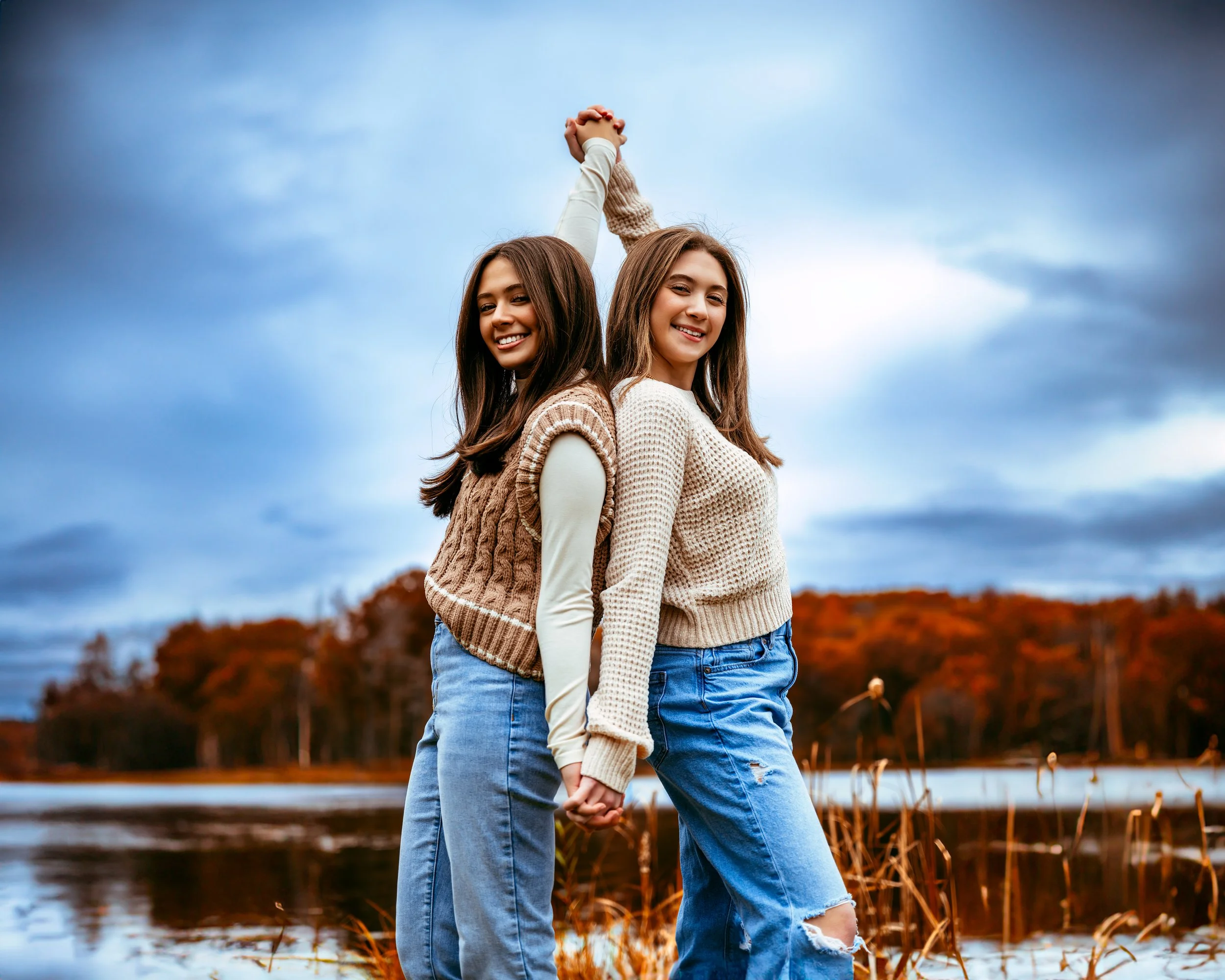 Two people smiling, holding hands with arms raised, standing in front of a lake with autumn trees in the background.