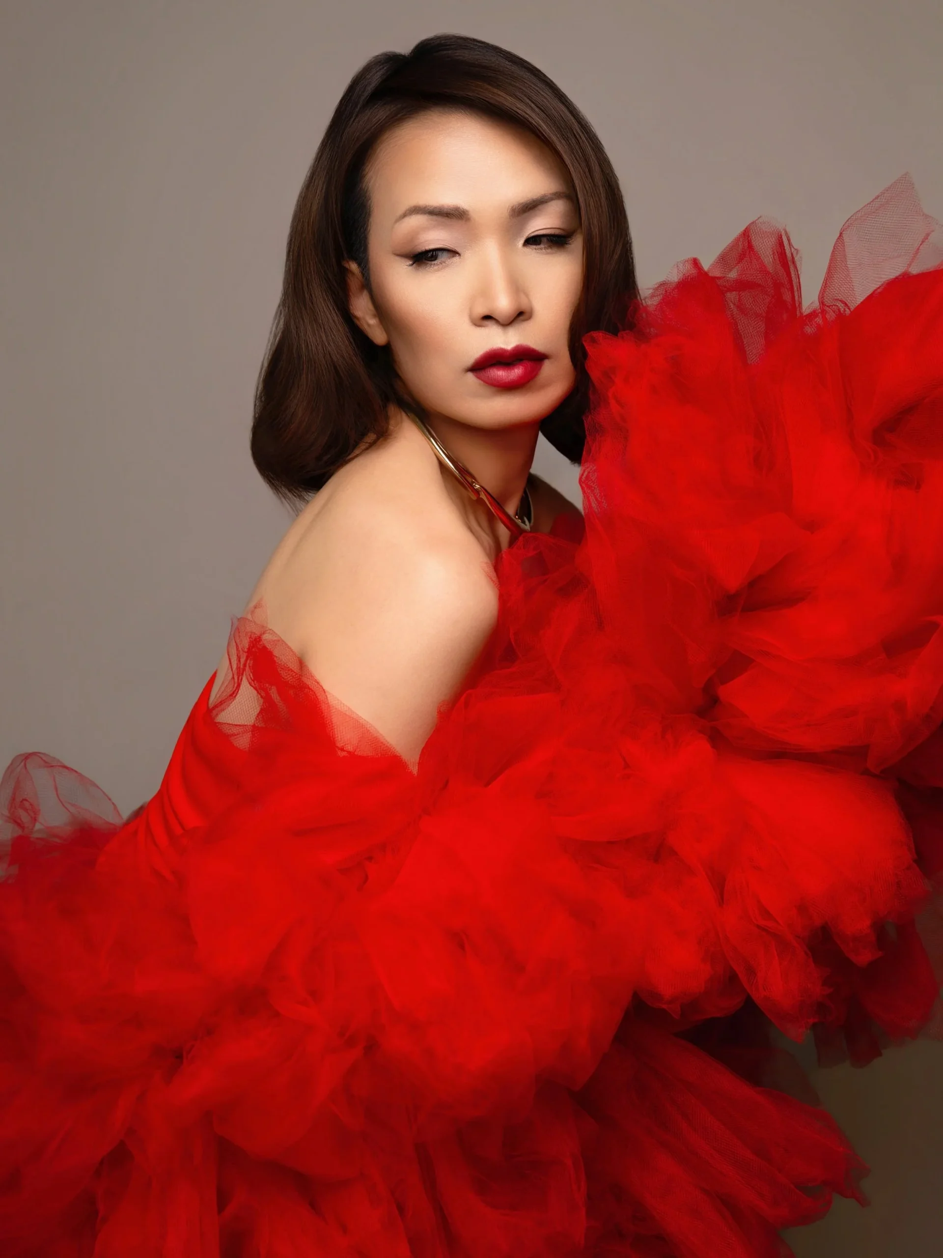 A woman with brown hair in loose waves, wearing red lipstick, and a red ruffled dress or costume with off-shoulder design, posed against a neutral gray background.