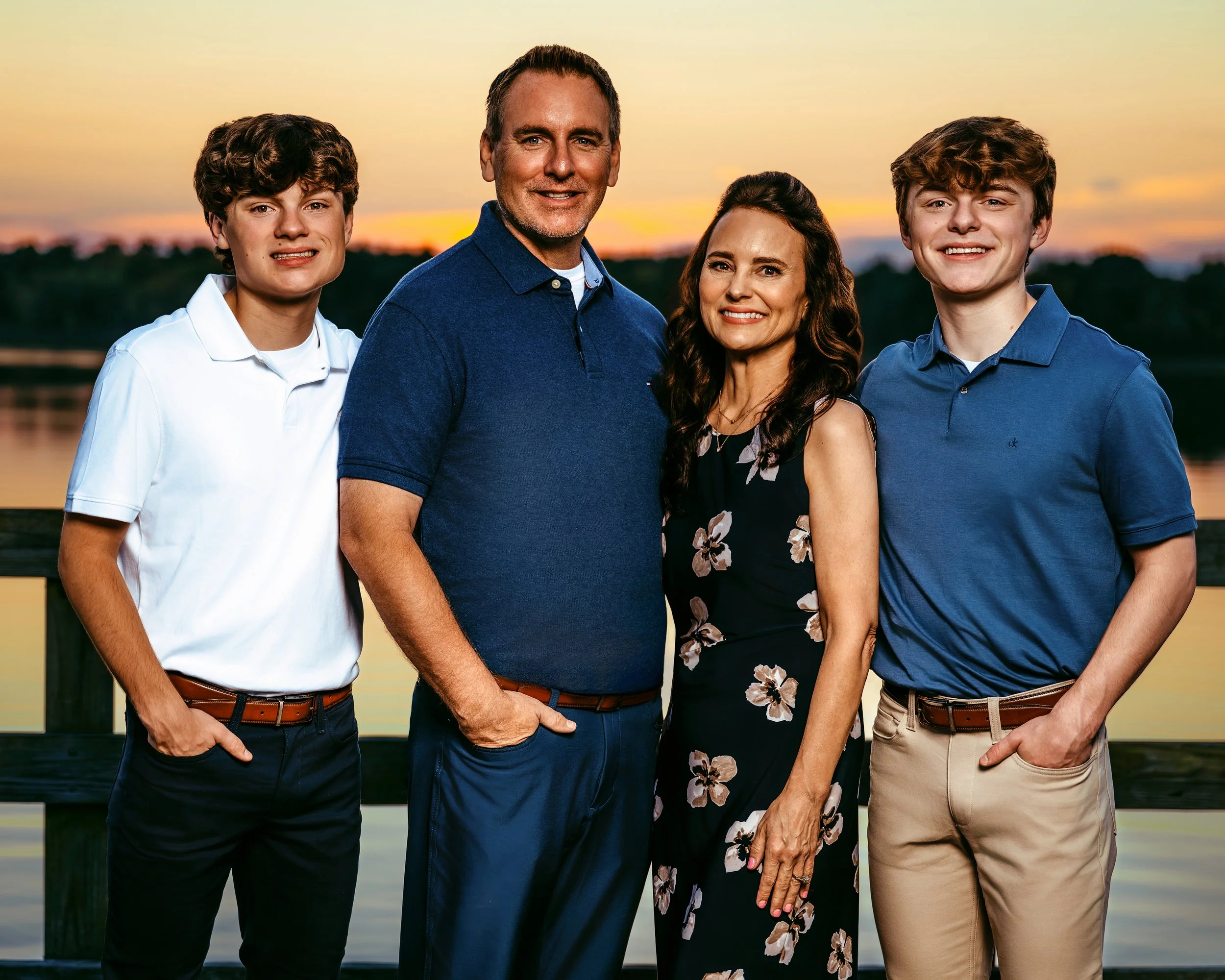 A family of four posing outdoors during sunset, near a body of water. The two adults are standing in the center, with the male wearing a navy blue shirt and the female in a floral dress. They are flanked by two young males in collared shirts.