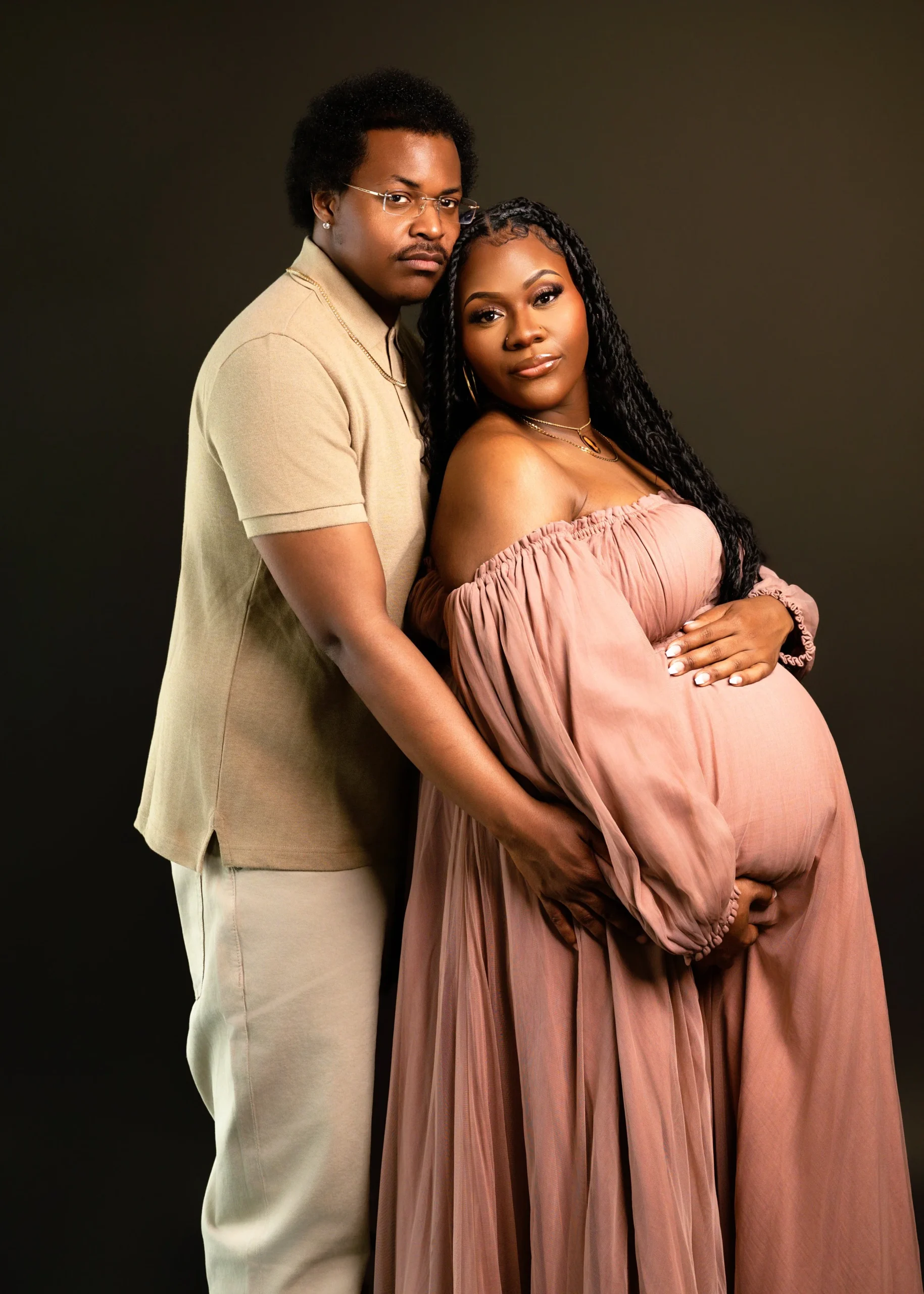 A couple, a man and a pregnant woman, standing close together against a dark background. The woman is wearing a long, off-shoulder peach-colored dress and the man is wearing a beige shirt and pants. They are posing affectionately with the man's arm a