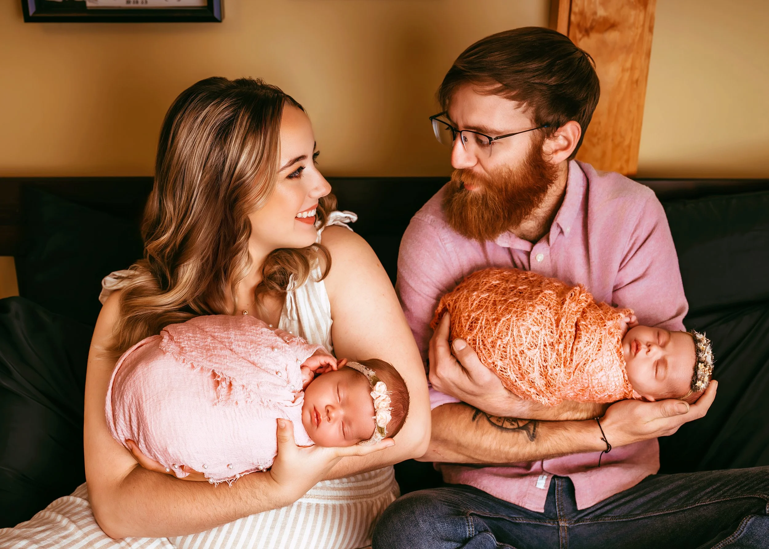 A couple holding their newborn babies, smiling and looking at each other in a cozy indoor setting.