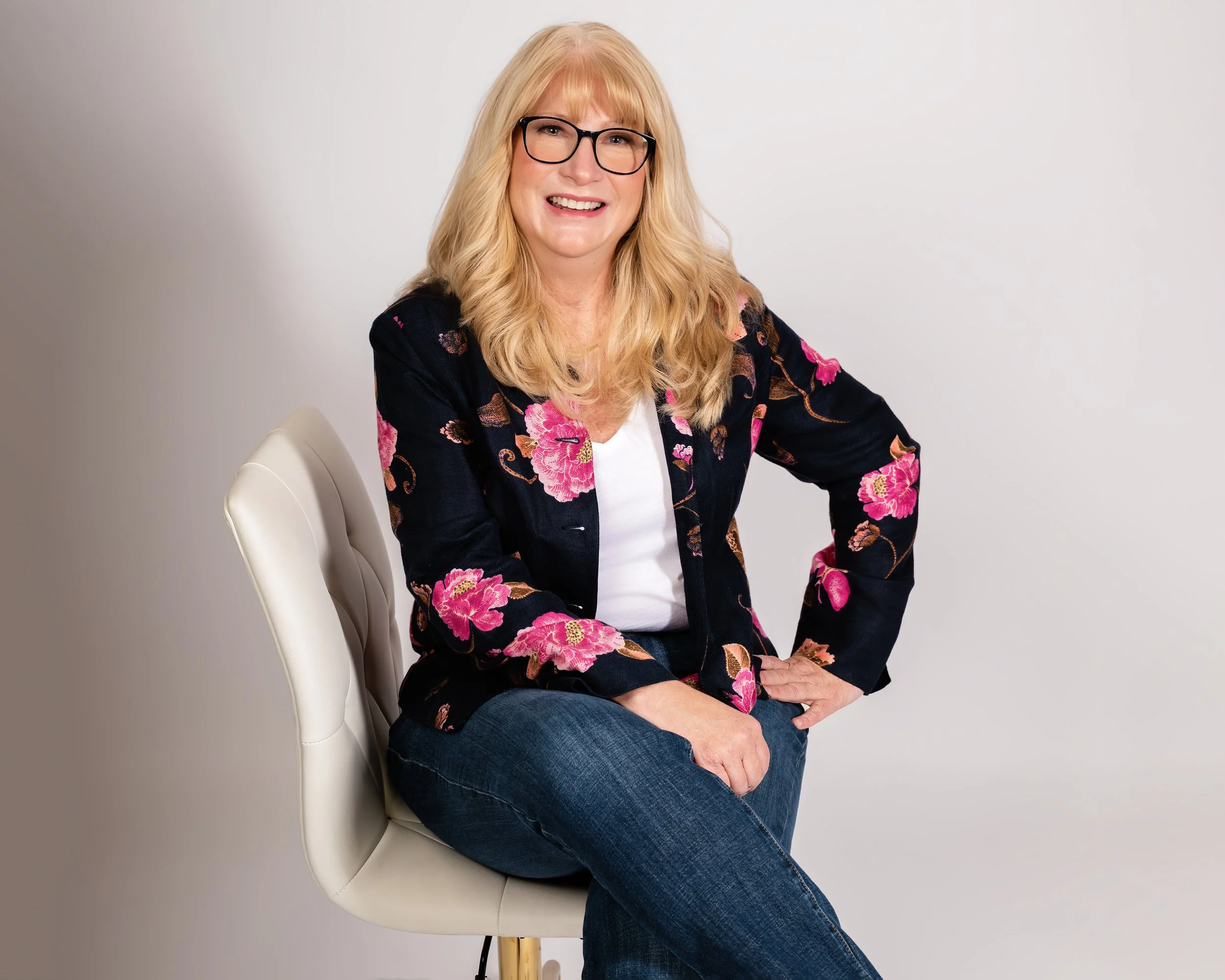 A woman with long blonde hair and glasses is sitting on a white chair. She is wearing a black blazer with pink floral patterns, a white top, and blue jeans. She is smiling and posed casually against a plain background.