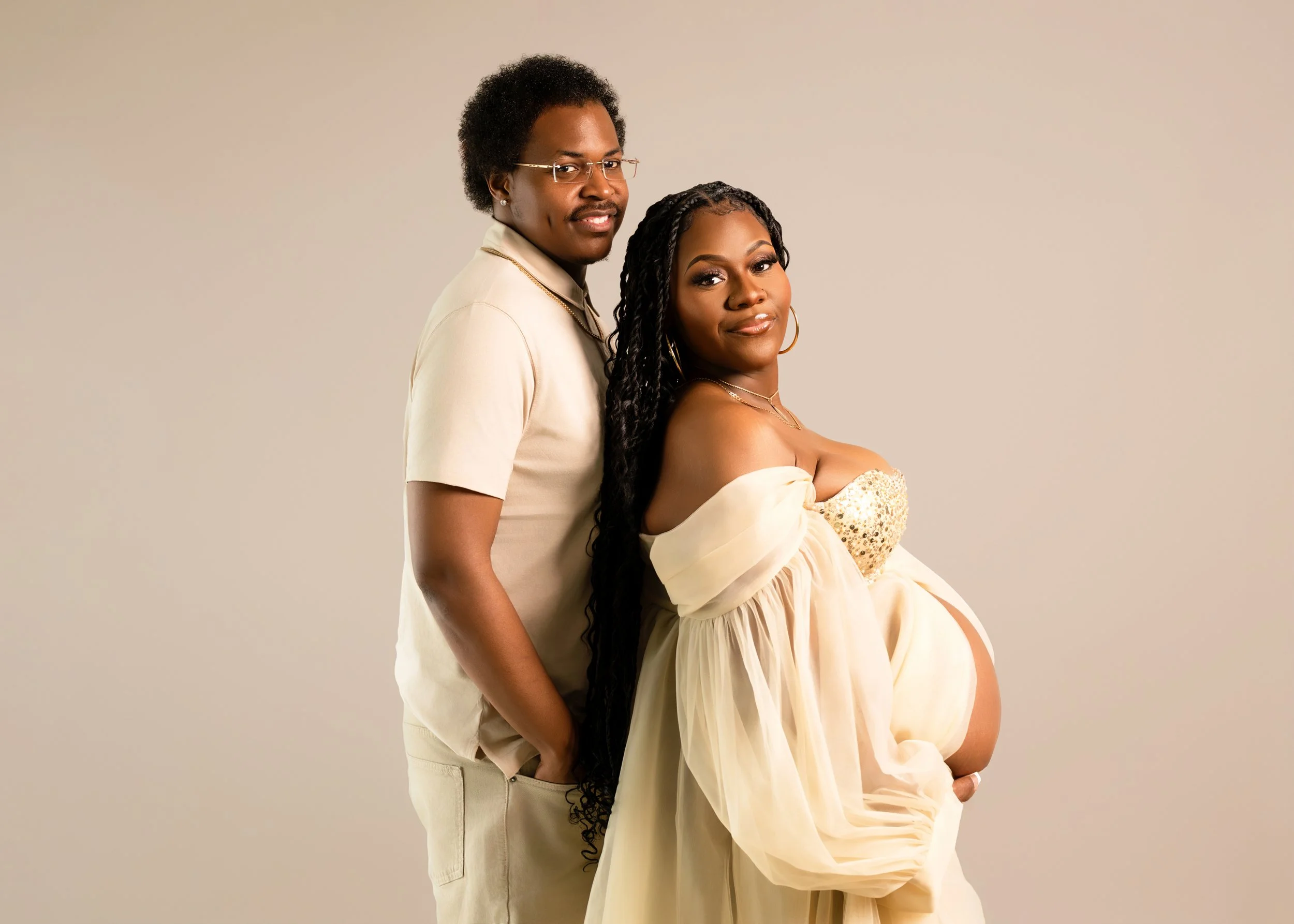 A pregnant woman in an off-the-shoulder gold-sequined dress with a sheer, flowing cream-colored gown, standing close to a man in a light beige shirt and matching pants, against a plain light-colored background.