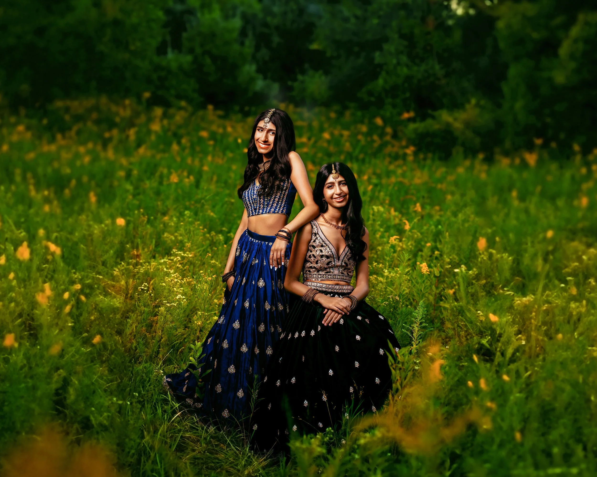 Two women in traditional Indian attire, standing and sitting in a field with green grass and yellow wildflowers.