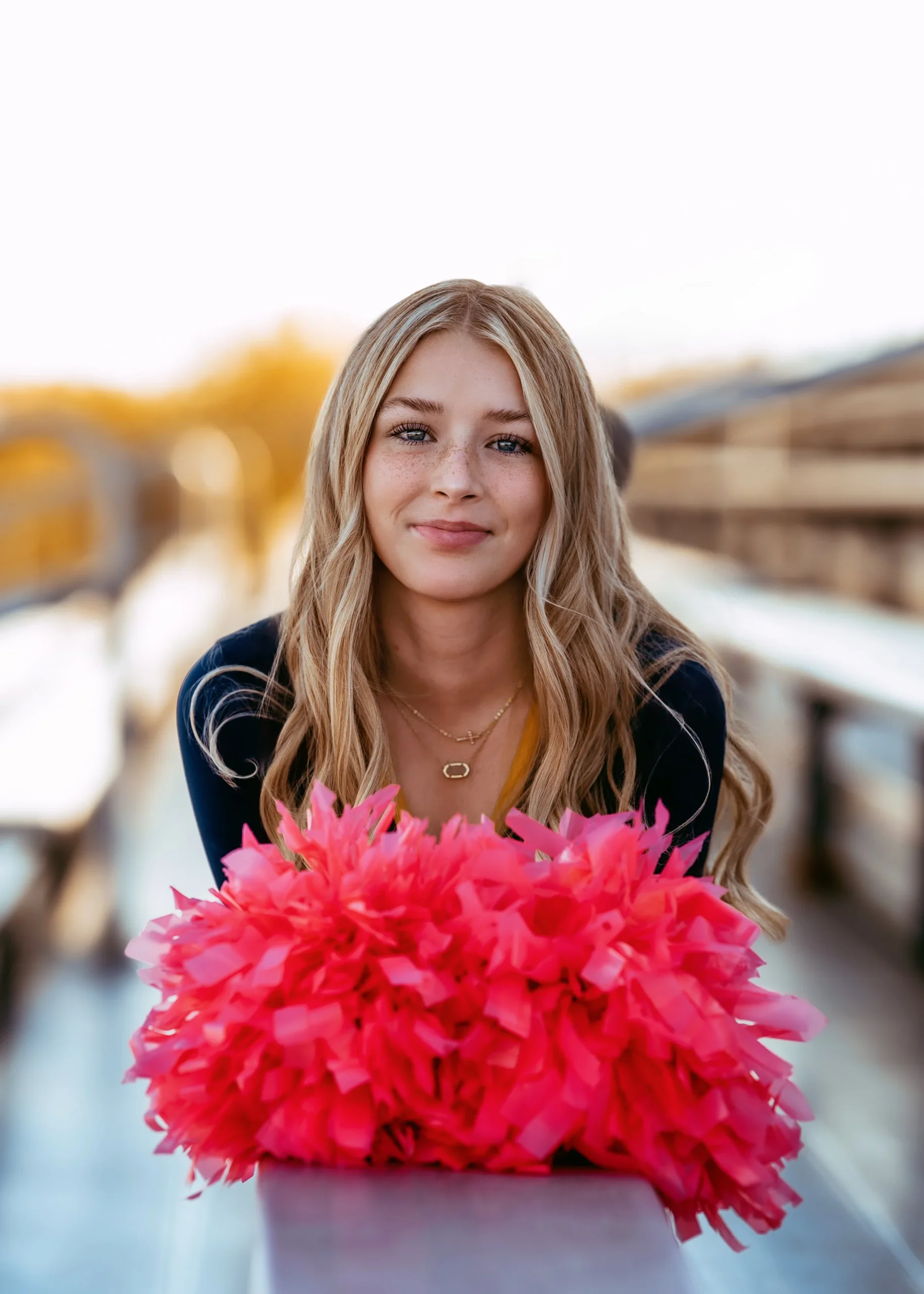A woman with long blonde hair holding hot pink pom-poms, wearing a dark top, sitting on bleachers outdoors with a blurred background.