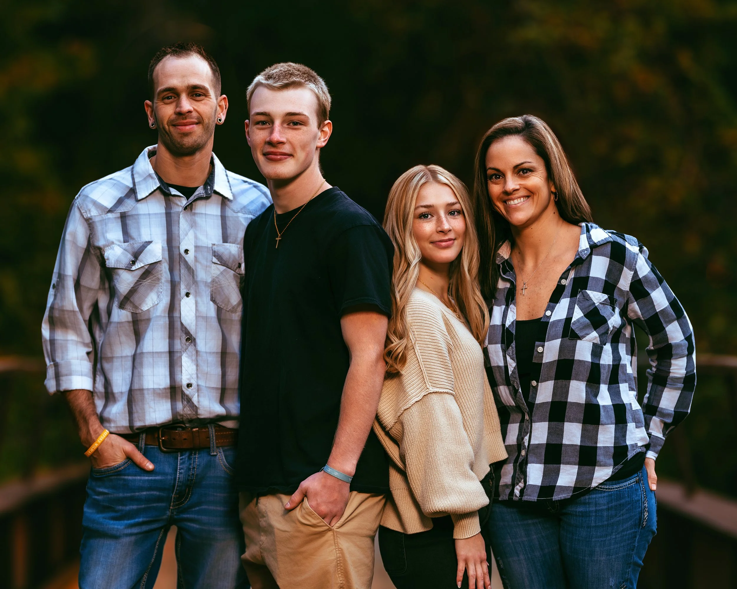 A group of four people outdoors, two men and two women, smiling and standing close together. The men are wearing casual shirts and jeans, while the women are wearing casual tops and jeans. The background is a blurred natural setting.