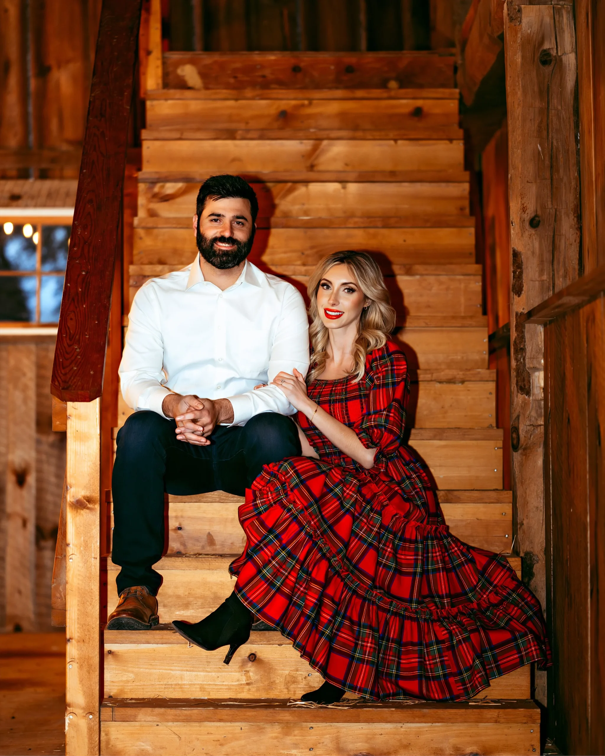 A couple sitting on wooden stairs, with the man in a white shirt and dark pants, and the woman in a red plaid dress.