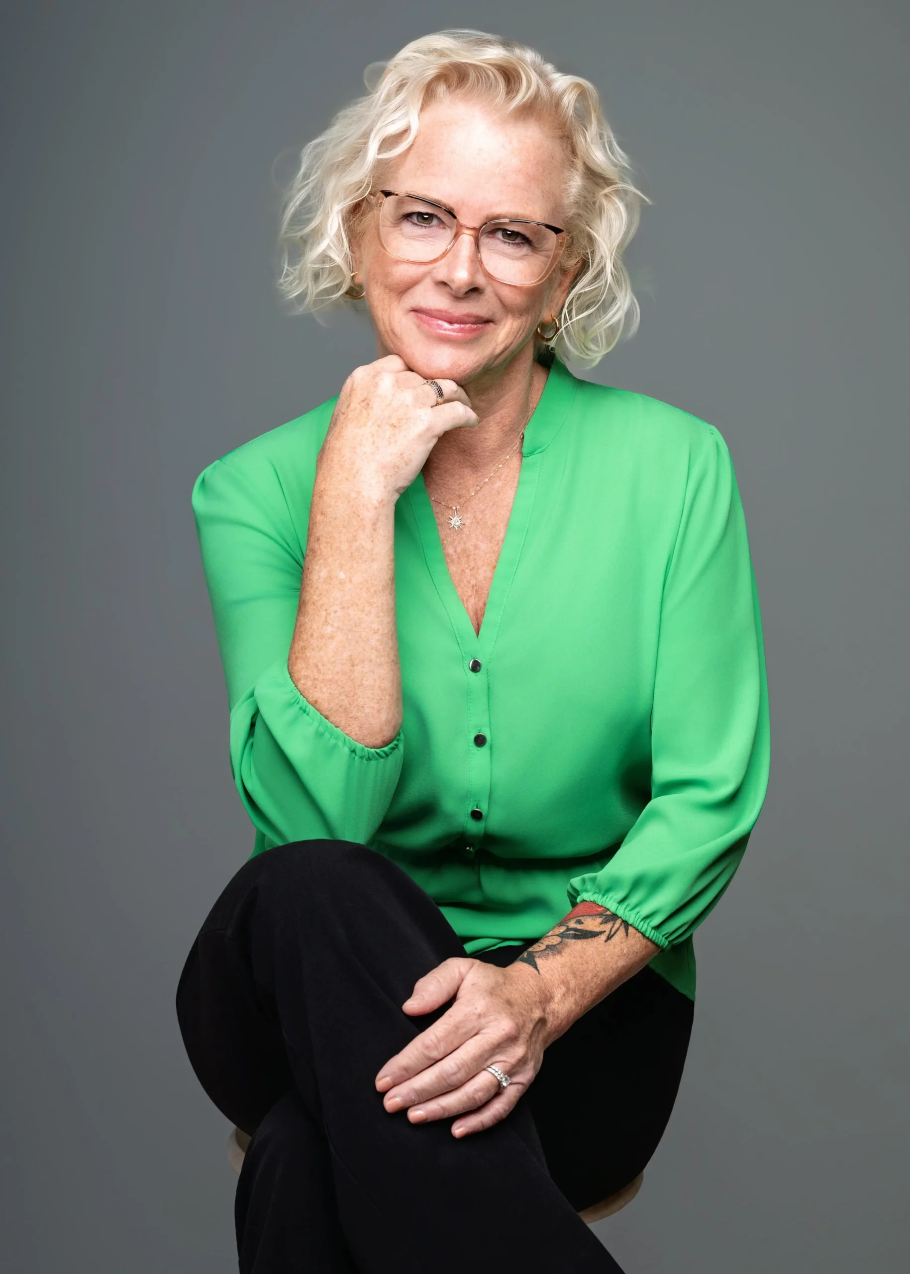 A woman with curly white hair, glasses, and a green blouse sitting against a gray background, smiling and resting her chin on her hand.
