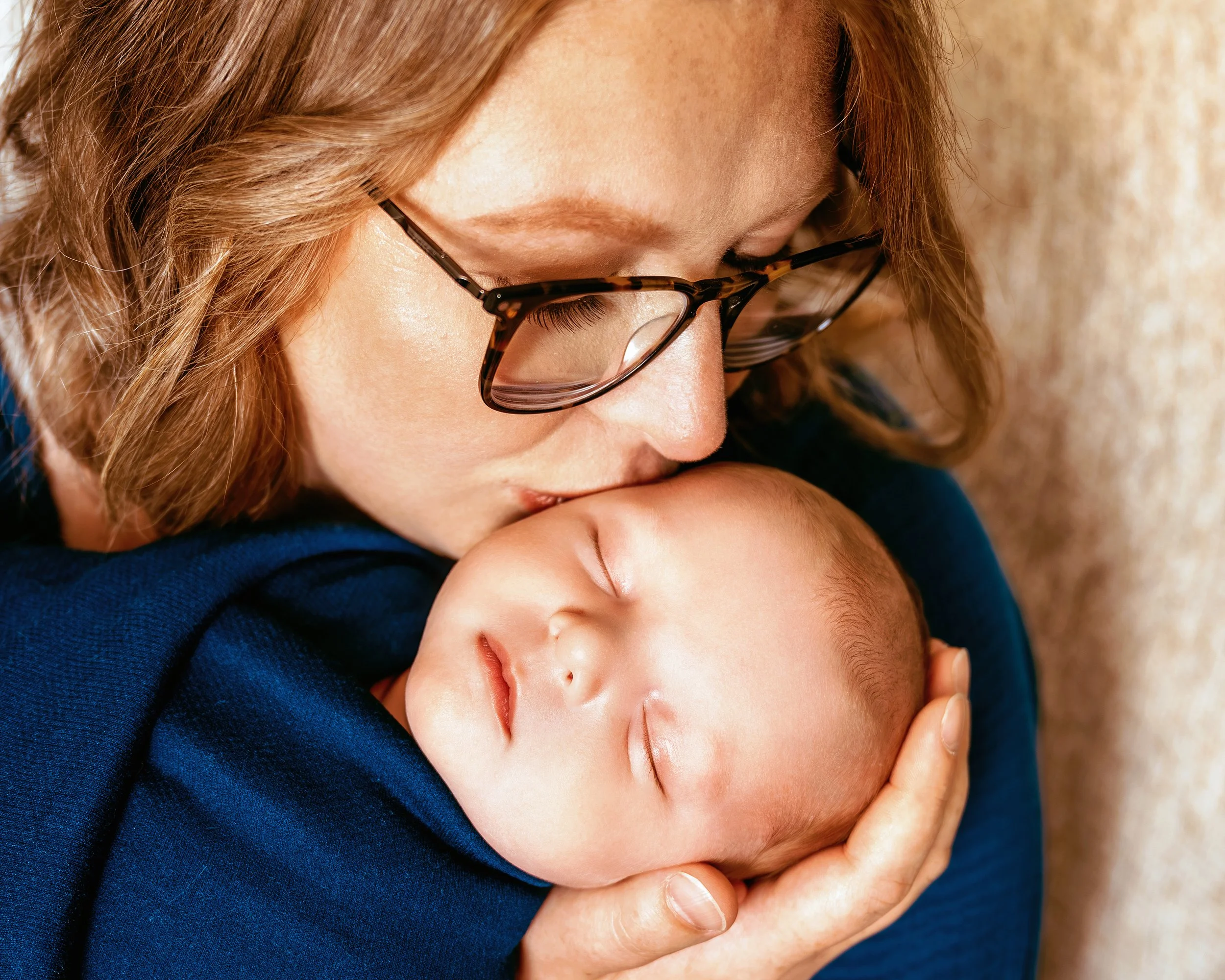 Close-up of a woman with glasses gently holding and kissing a sleeping baby.