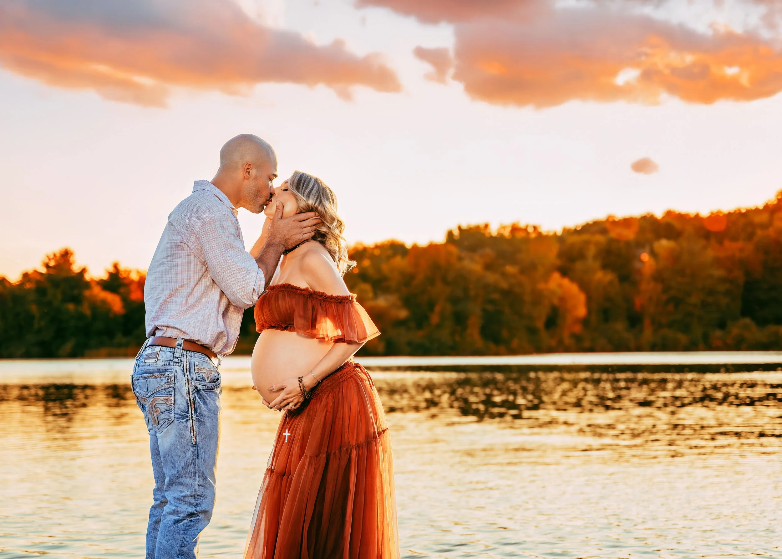 A pregnant woman in a red dress and a man are sharing a kiss by a lake at sunset, with trees in the background.