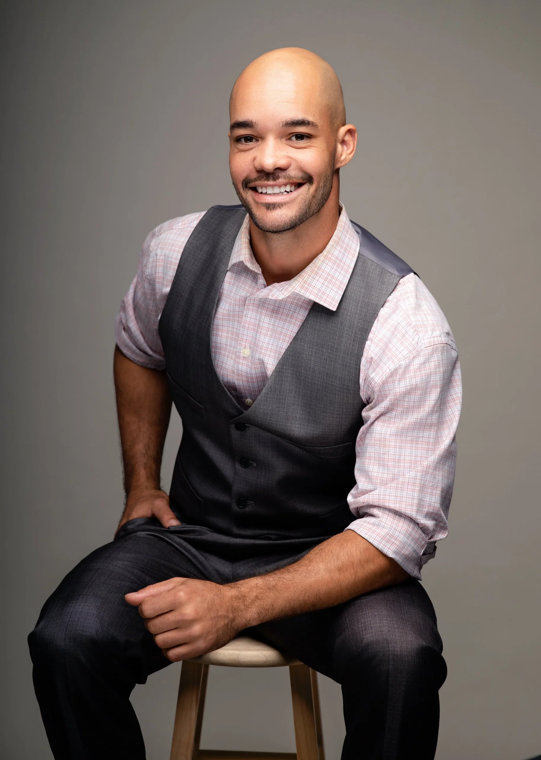 A smiling man with a bald head, wearing a pink checkered shirt and a gray vest, sitting on a stool against a plain gray background.