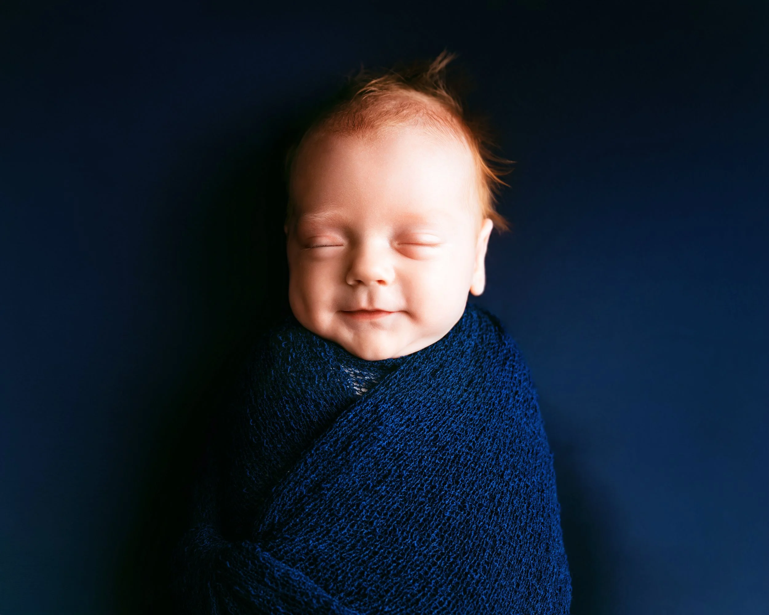 Young smiling baby with closed eyes wrapped in a navy blue knit blanket against a matching dark blue background.