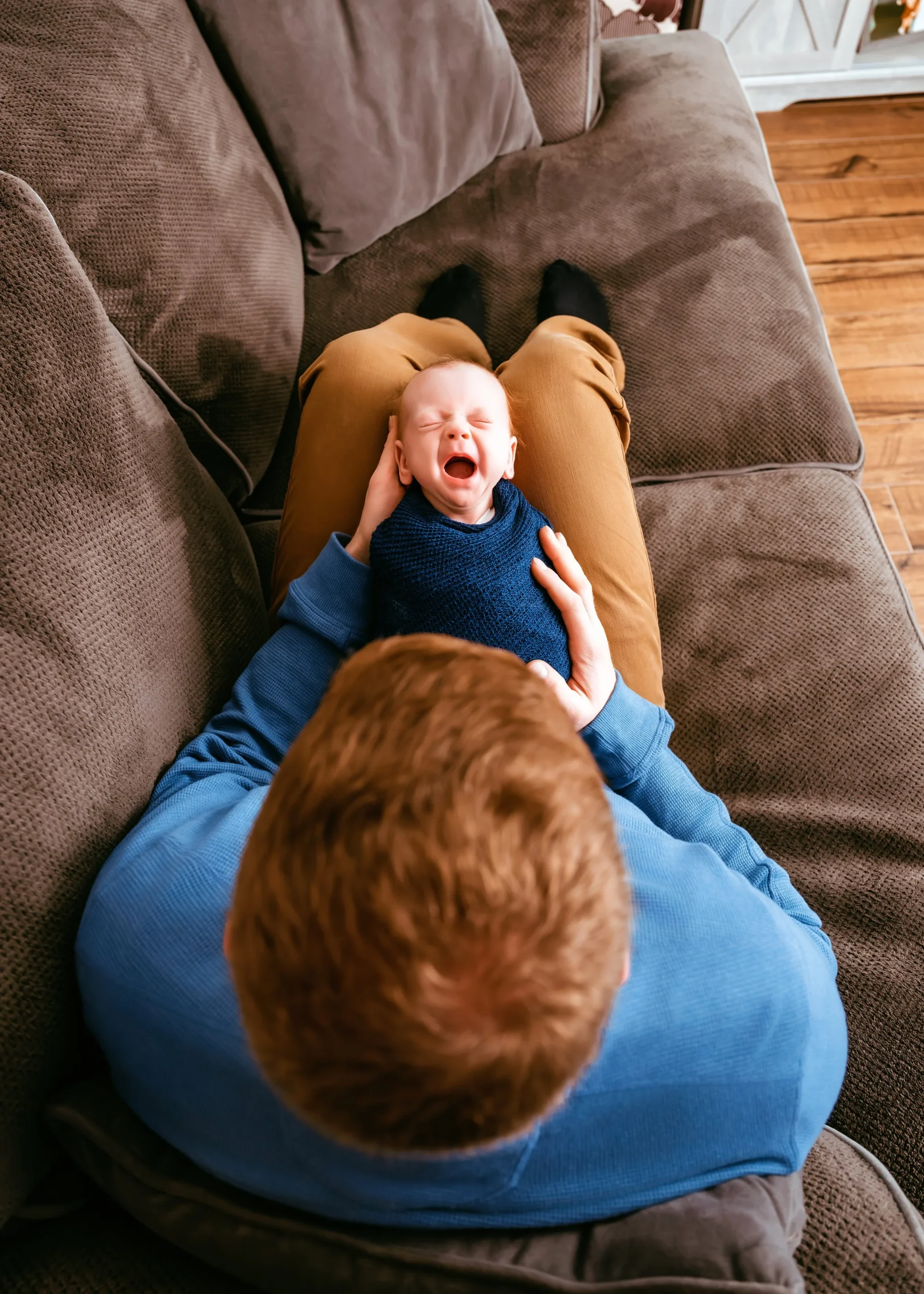 Person sitting on a couch holding a yawning baby wrapped in a blue blanket.