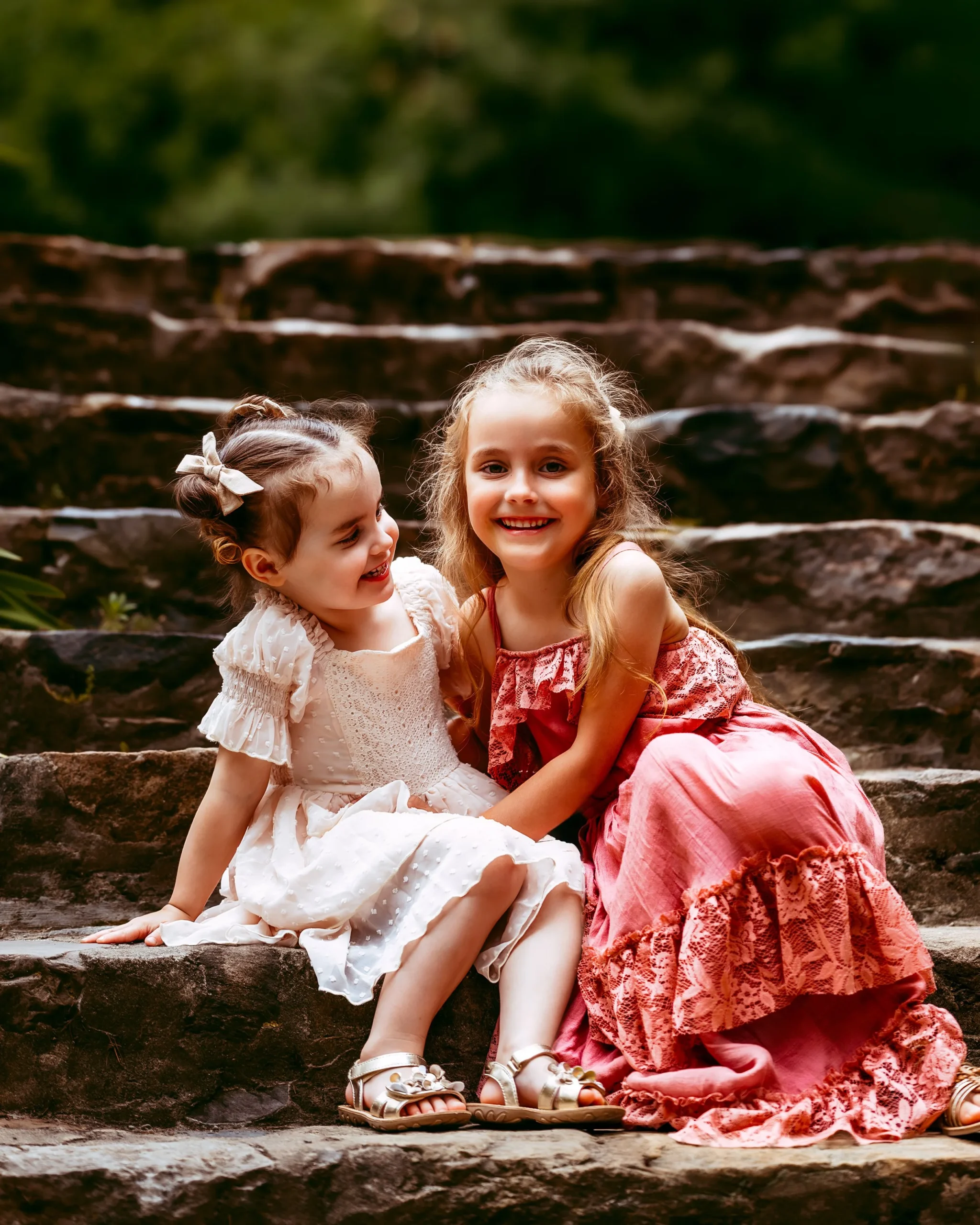 Two young girls in elegant dresses sitting on stone steps, smiling and looking at each other.