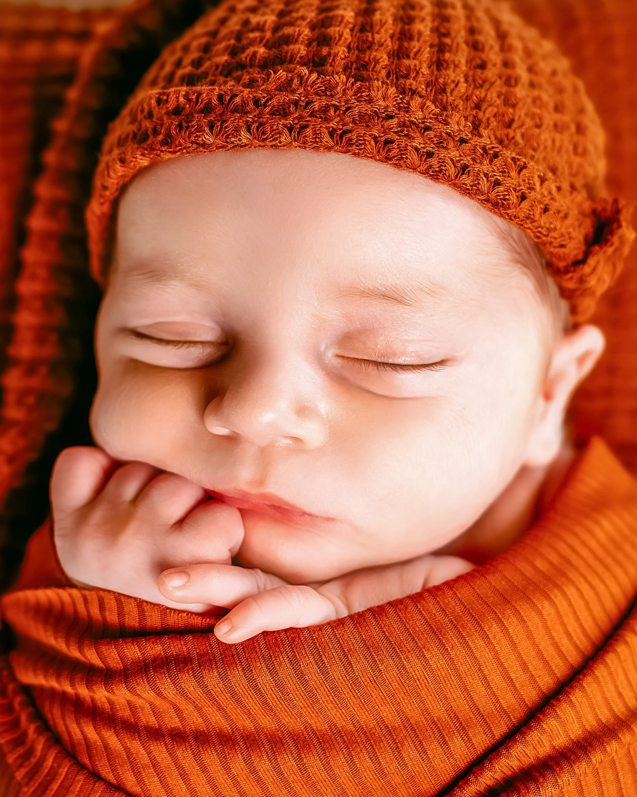 Close-up of a sleeping newborn baby wearing an orange knitted hat and swaddled in an orange blanket.
