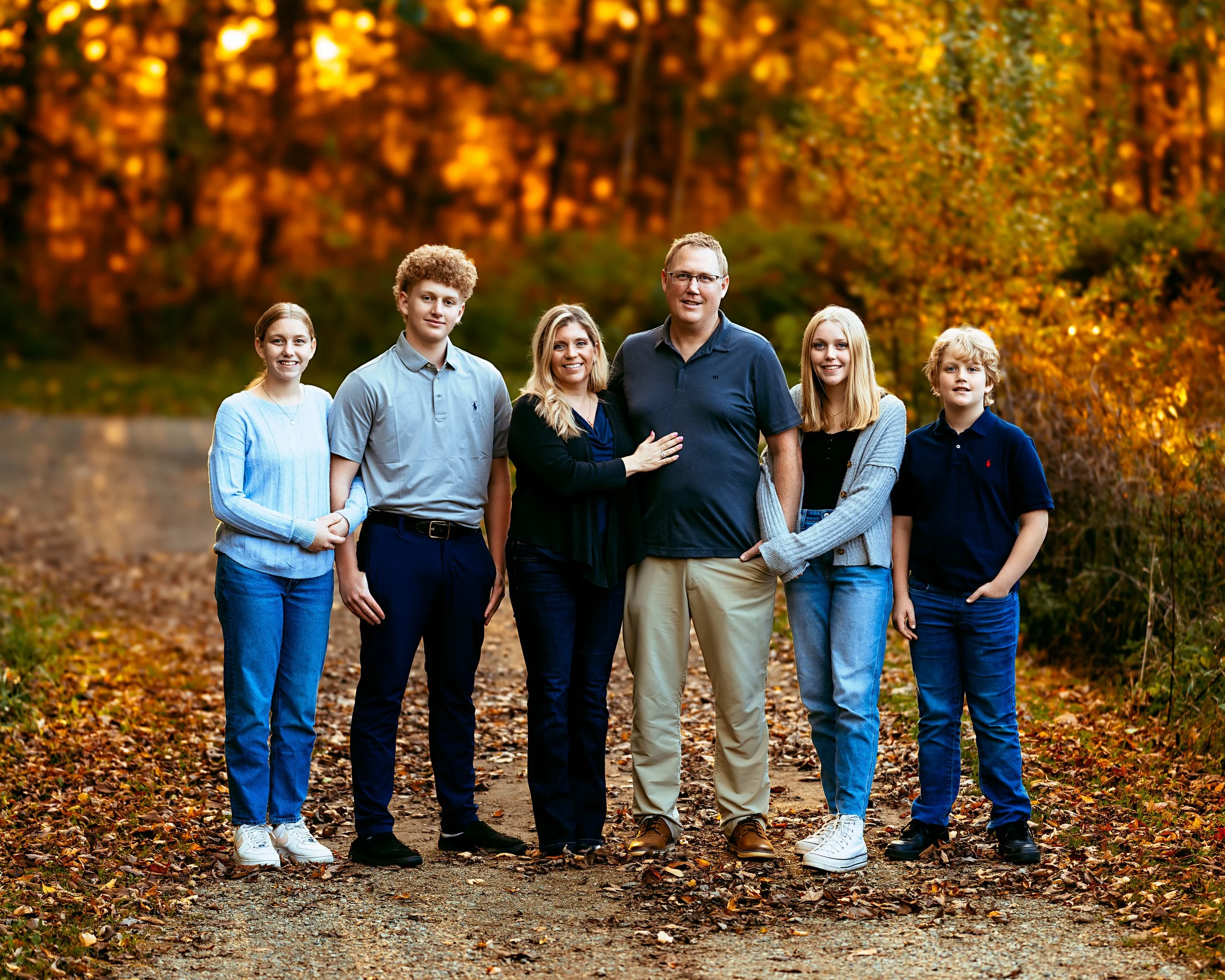 A family of six posing for a photo in a forest setting with autumn foliage. The group includes three males and three females, all standing closely together on a leaf-covered path. The background features vibrant fall colors with orange and yellow lea