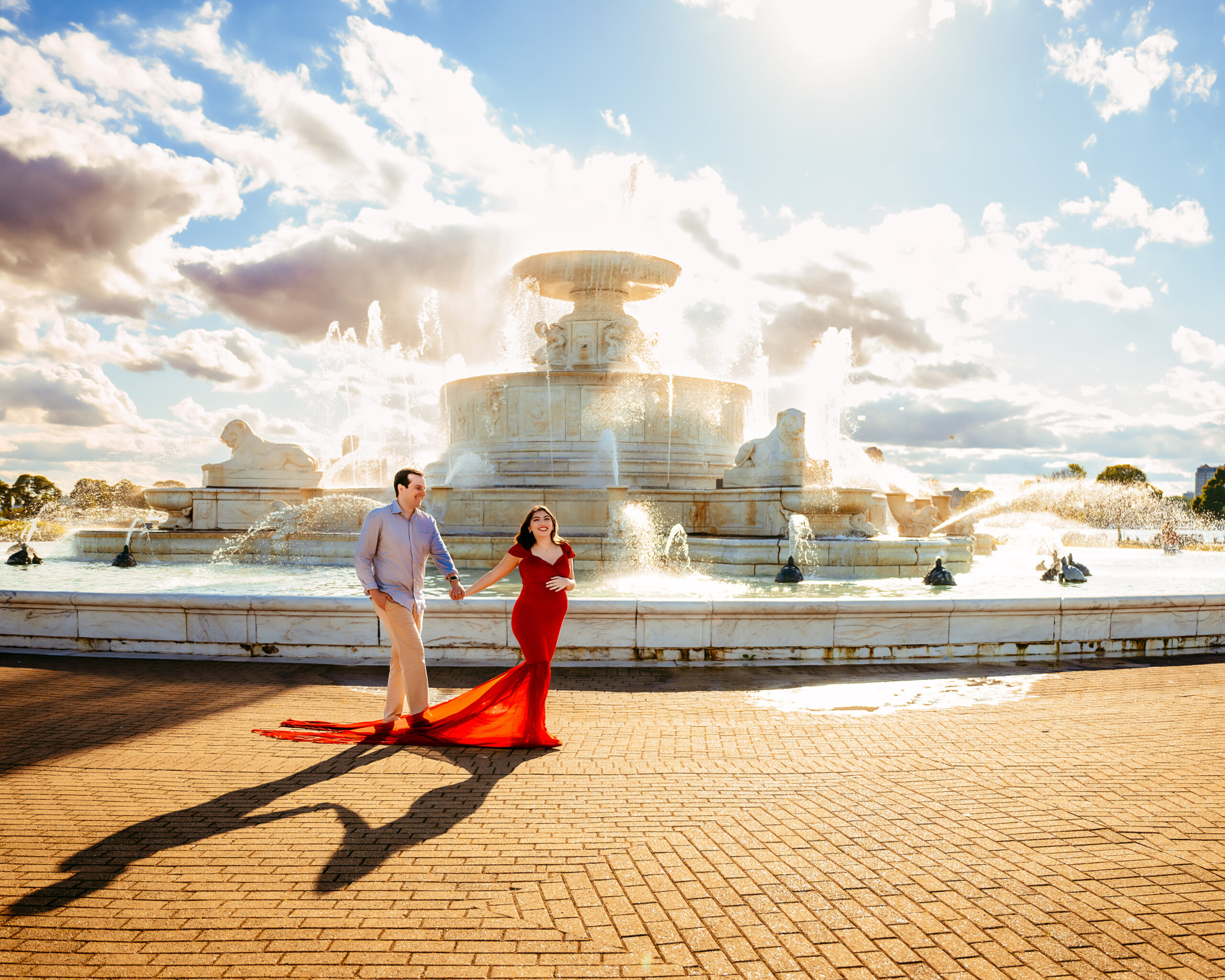 Couple holding hands and walking in front of a large ornate fountain, woman in a red gown and the man in a light shirt and beige pants, long shadows on a brick plaza.