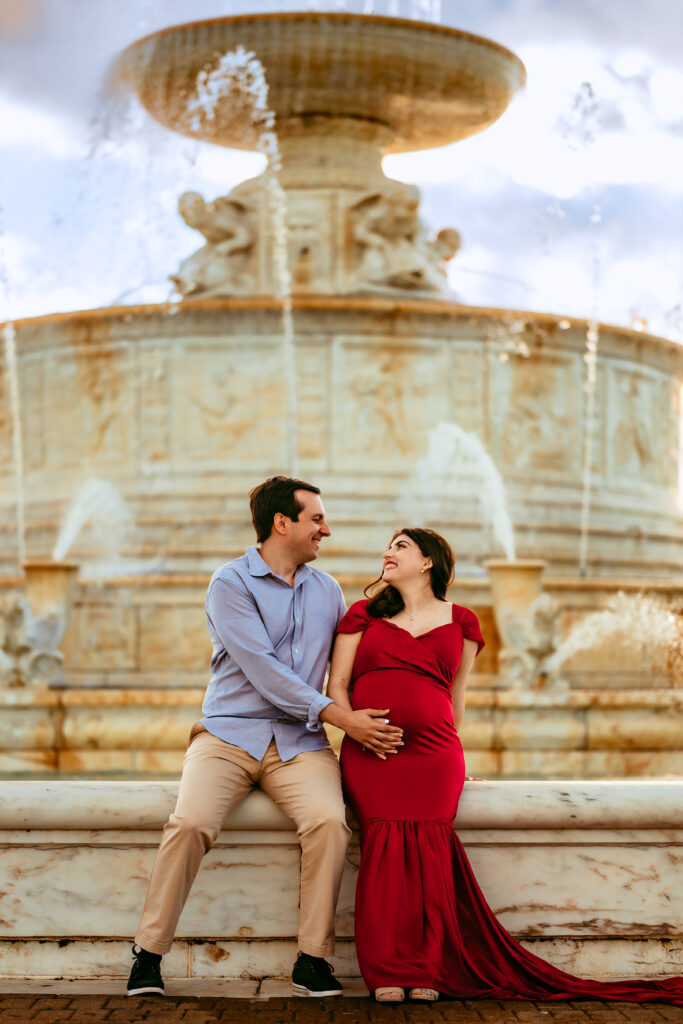 Pregnant woman in a red gown sits beside her partner on a fountain ledge, both smiling at each other with a grand fountain in the background.