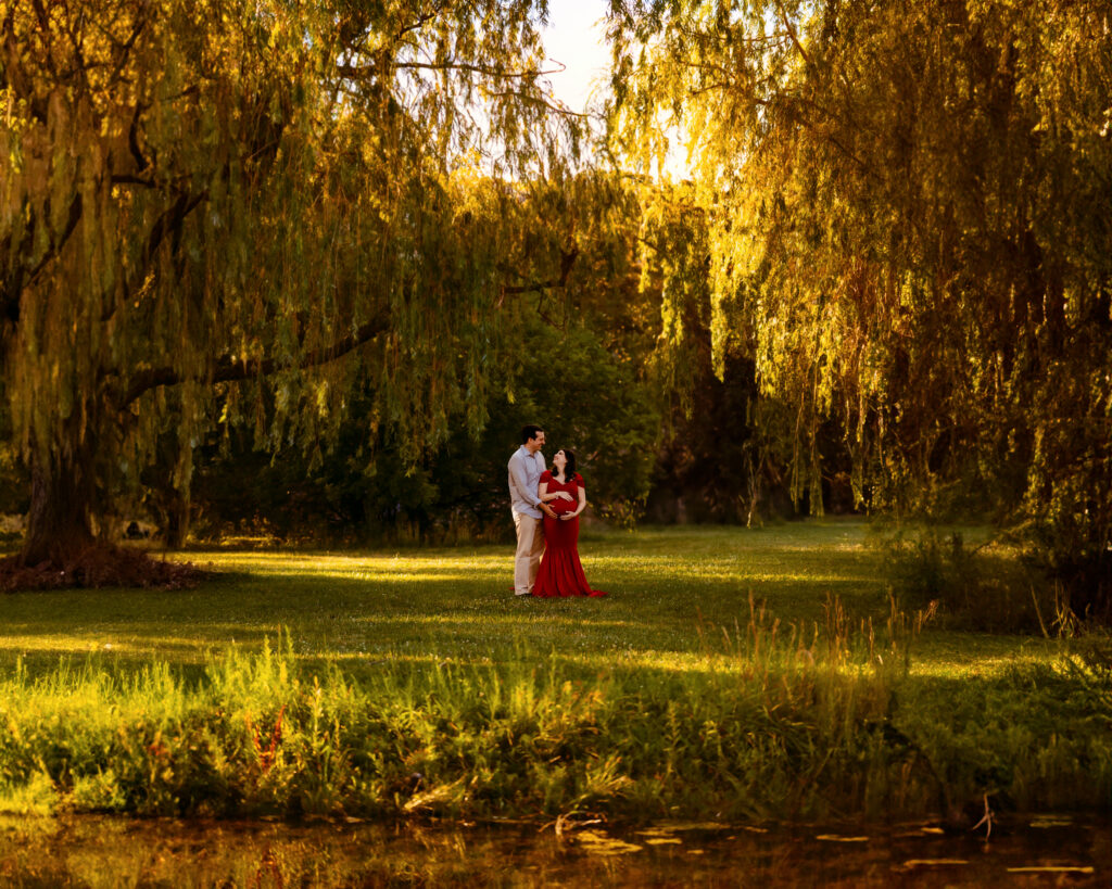 A couple in a red dress and a light shirt stand on a sunlit lawn under cascading golden willow branches in a park.