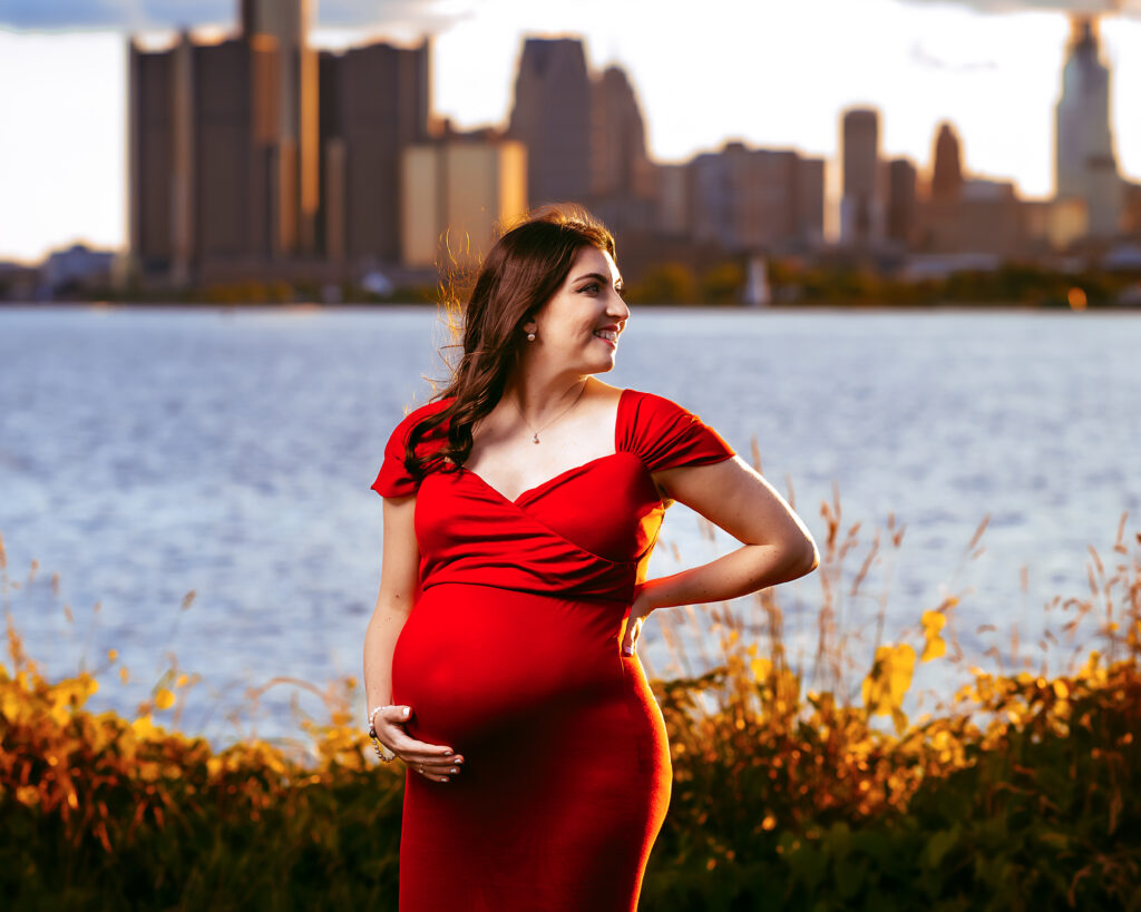 Pregnant woman in a red dress standing by a lake with a city skyline in the background at sunset.