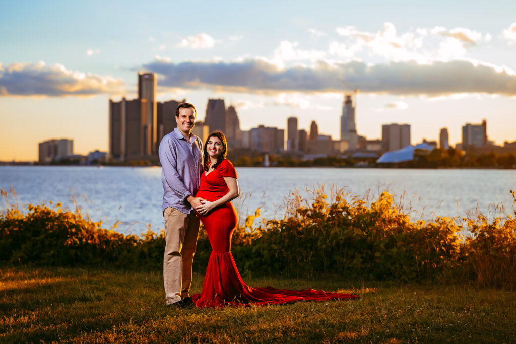 Pregnant woman in a red gown and her partner smile by a lake at sunset with a city skyline in the background.
