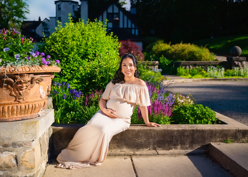 Aclipen_FINAL_5660 | Two Wild Souls Photography Woman in a peach off-shoulder dress sits on stone steps in a colorful garden full of green shrubs and purple flowers, smiling at the camera.