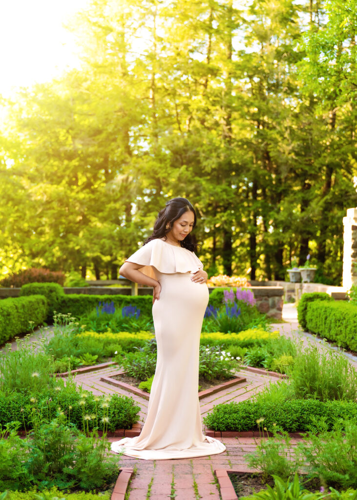Aclipen_FINAL_5710 | Two Wild Souls Photography Pregnant woman in a pale pink off-the-shoulder gown standing in a sunlit garden, hands resting on her baby bump.