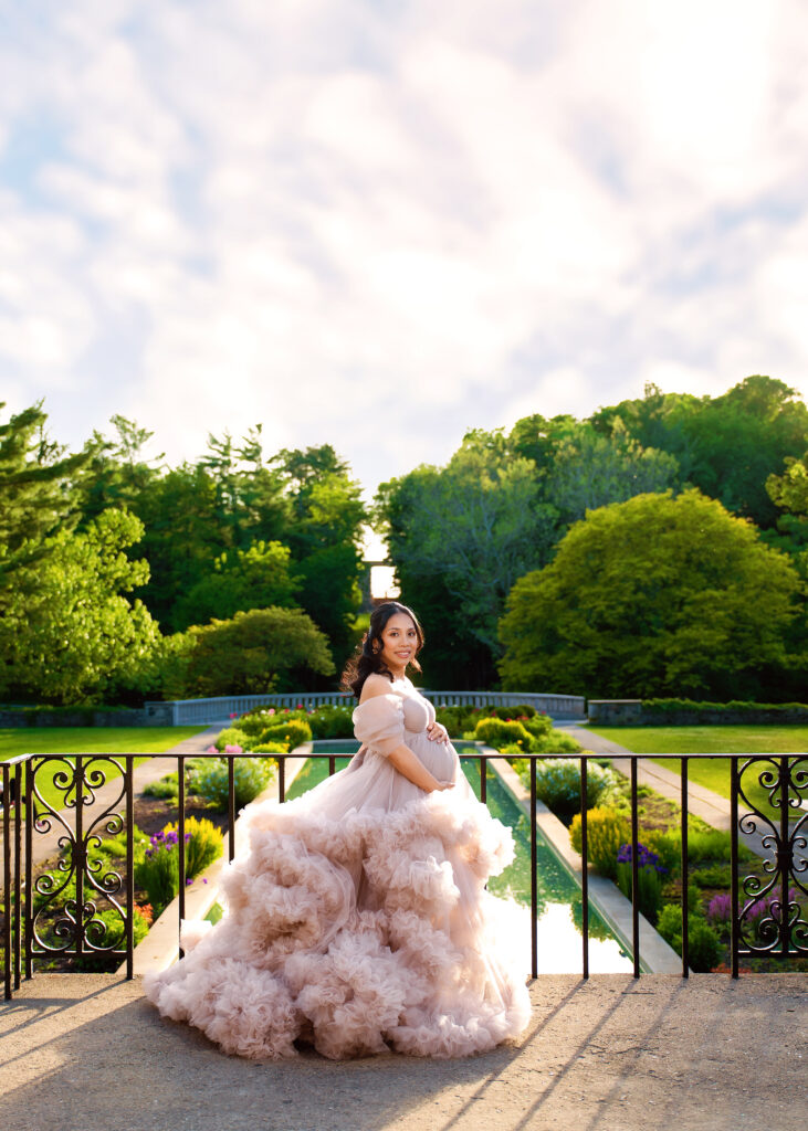 Aclipen_FINAL_5829 | Two Wild Souls Photography Pregnant woman in a voluminous pink gown posing on a decorative bridge in a lush garden setting.