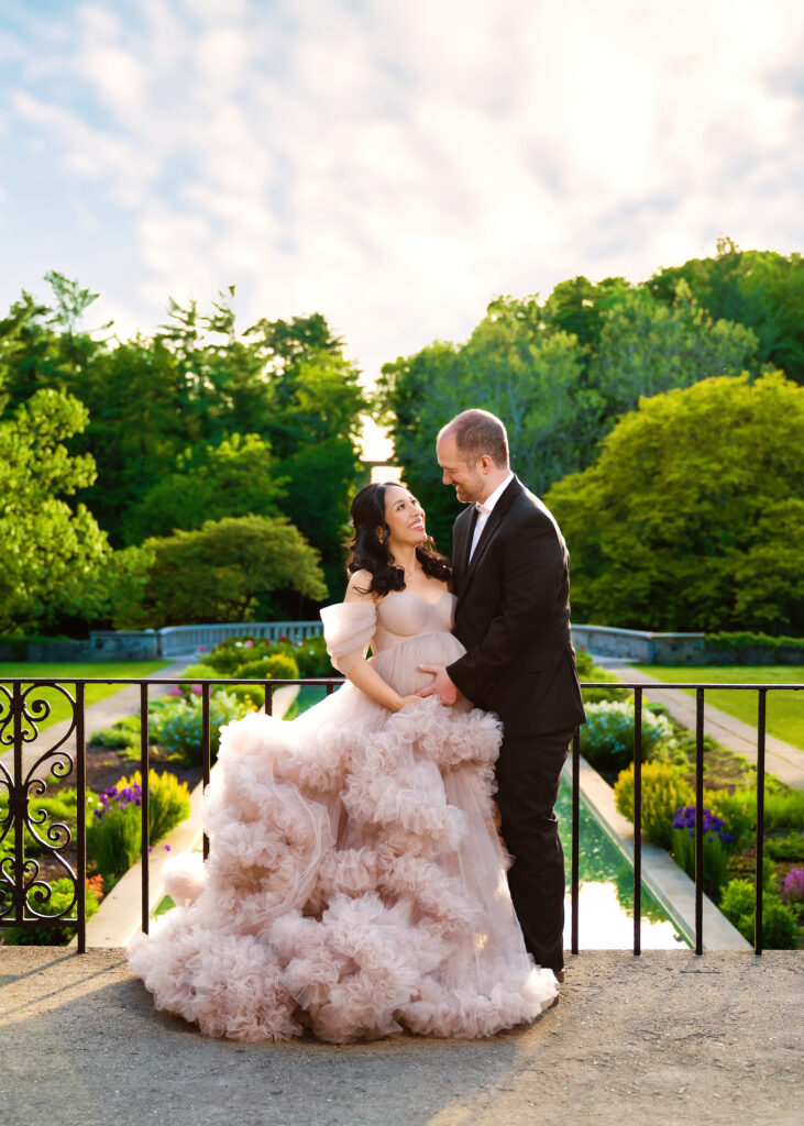 Aclipen_FINAL_5847 | Two Wild Souls Photography Pregnant bride in a blush pink, ruffled gown with her partner in a suit, smiling at each other in a sunny garden.