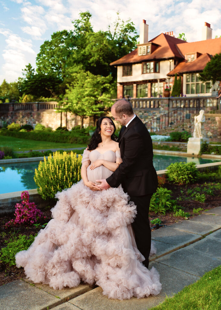Aclipen_FINAL_5897 | Two Wild Souls Photography A pregnant woman in a pale pink, ruffled gown smiling as a man in a suit embraces her outdoors by a garden and villa backdrop.