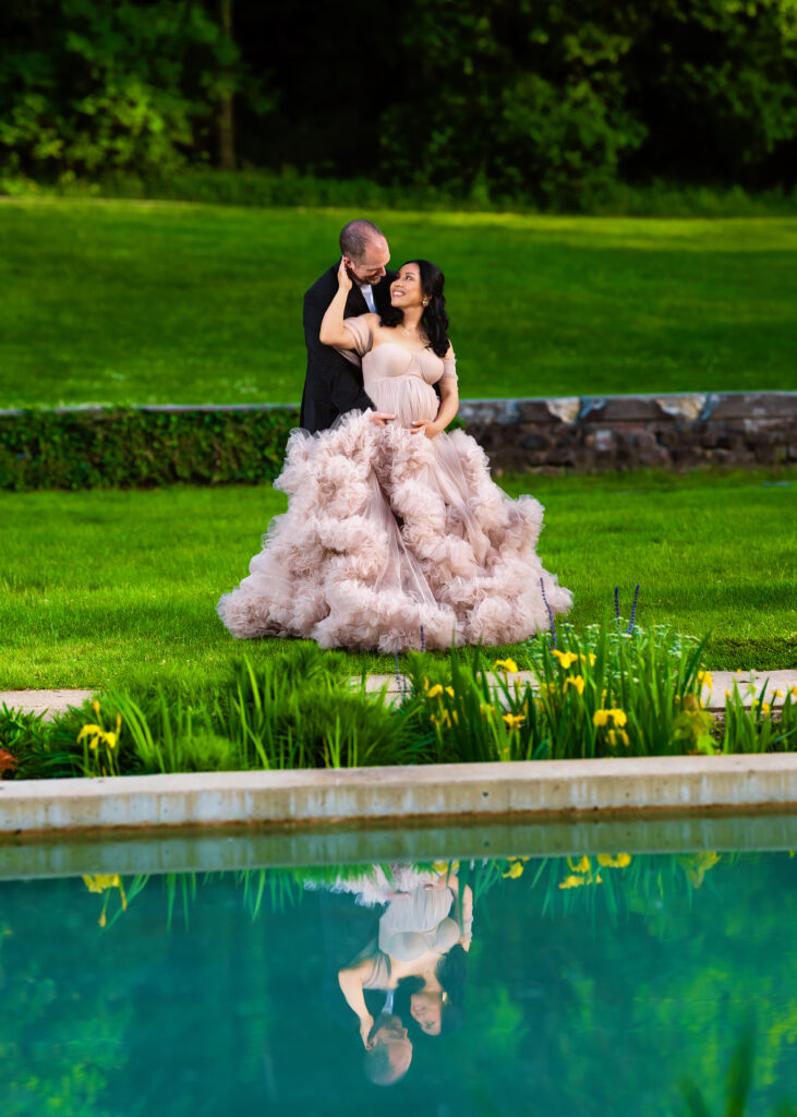 Aclipen_FINAL_5925 | Two Wild Souls Photography Pregnant woman in a pink ruffled gown with a man in a dark suit embracing in a park by a pond, their reflection visible in the water.