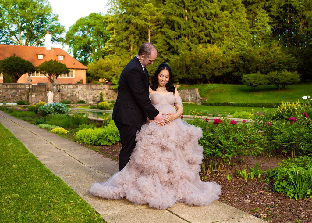 Aclipen_FINAL_5990 | Two Wild Souls Photography A couple outdoors in a garden: a man in a black suit gently holding a pregnant woman in a pale pink, ruffled ball gown while she smiles.