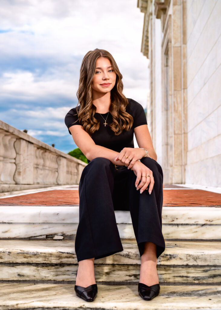 Woman in a black outfit sitting on marble steps outdoors, smiling at the camera, with a stone building beside her.