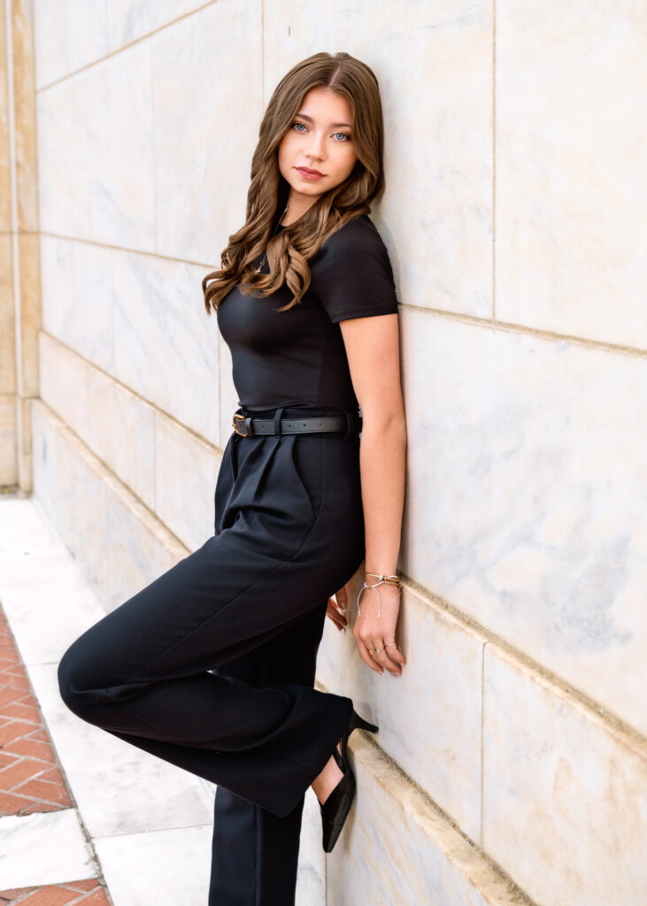 Woman in a black dress leaning against a pale stone wall, looking at the camera with a slight smile.