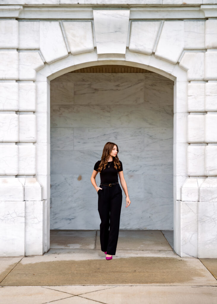 Woman in a black outfit stands in a marble arch doorway of a white stone building, wearing pink shoes and posing with hands in pockets.