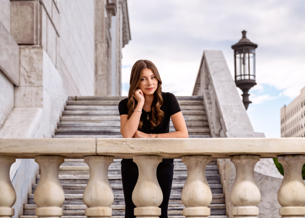 Woman in a black top rests her chin on her hand while leaning on a stone balustrade on a marble staircase outdoors.