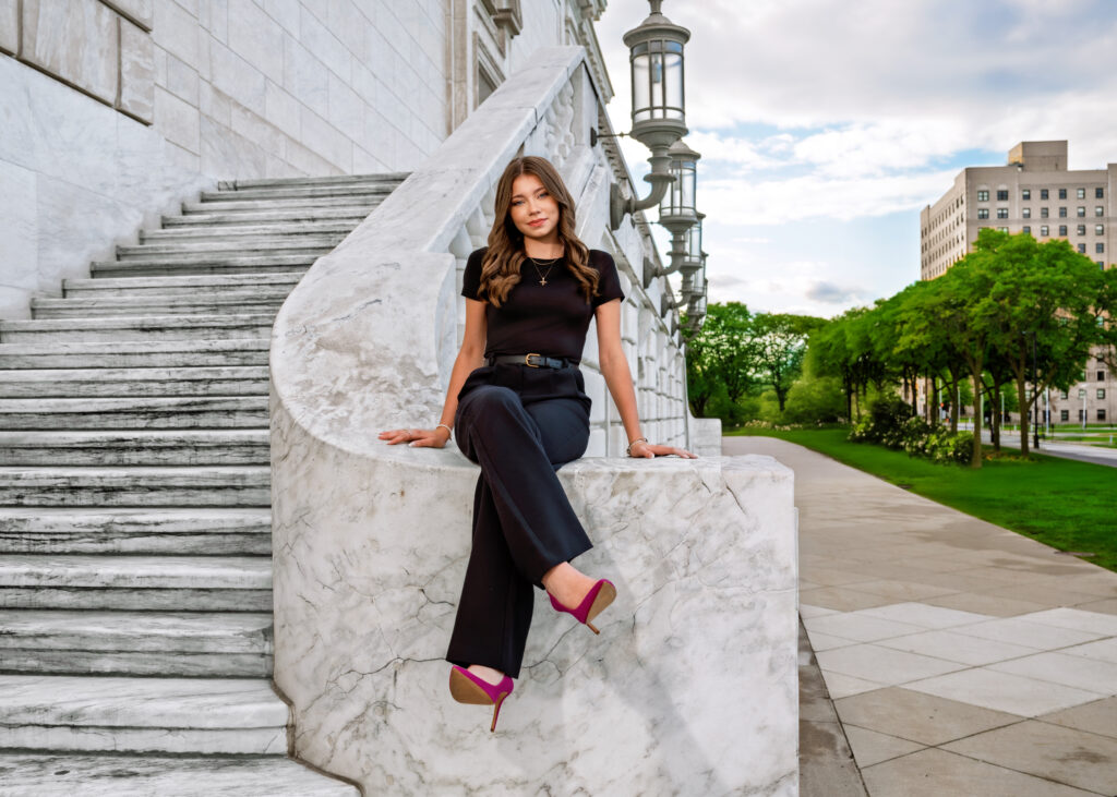 Woman in a black outfit sits on marble steps beside a historic stone railing with ornate lamps, urban park in the background.